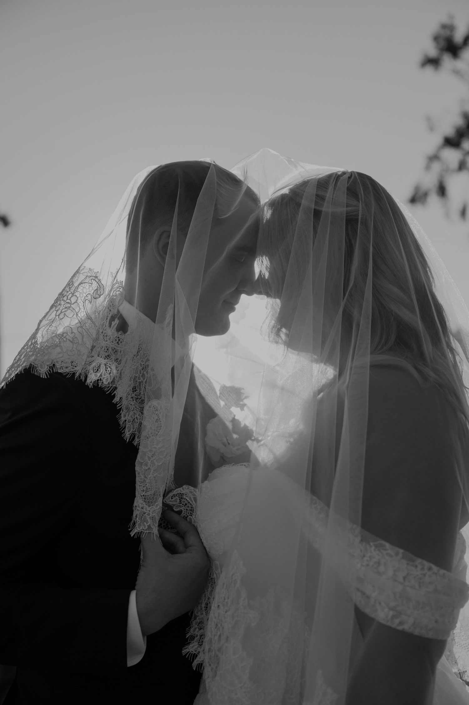 Black and white photo of bride and groom sharing intimate moment under lace wedding veil.