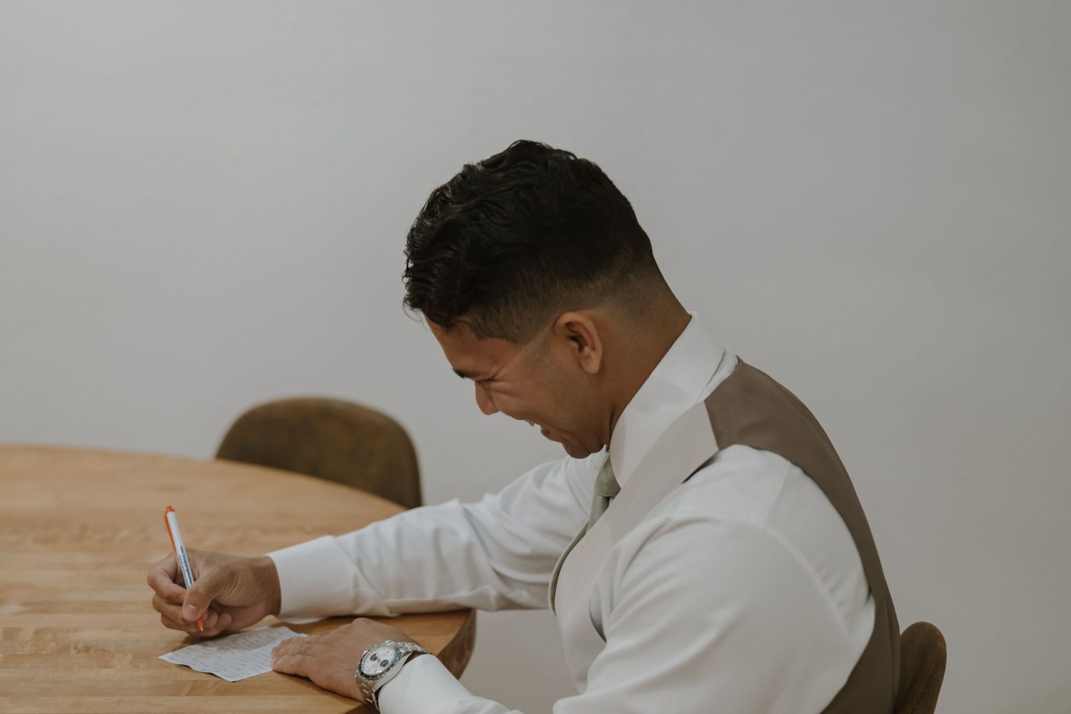 Groom in white shirt and vest writing heartfelt wedding vows at a wooden table before ceremony.