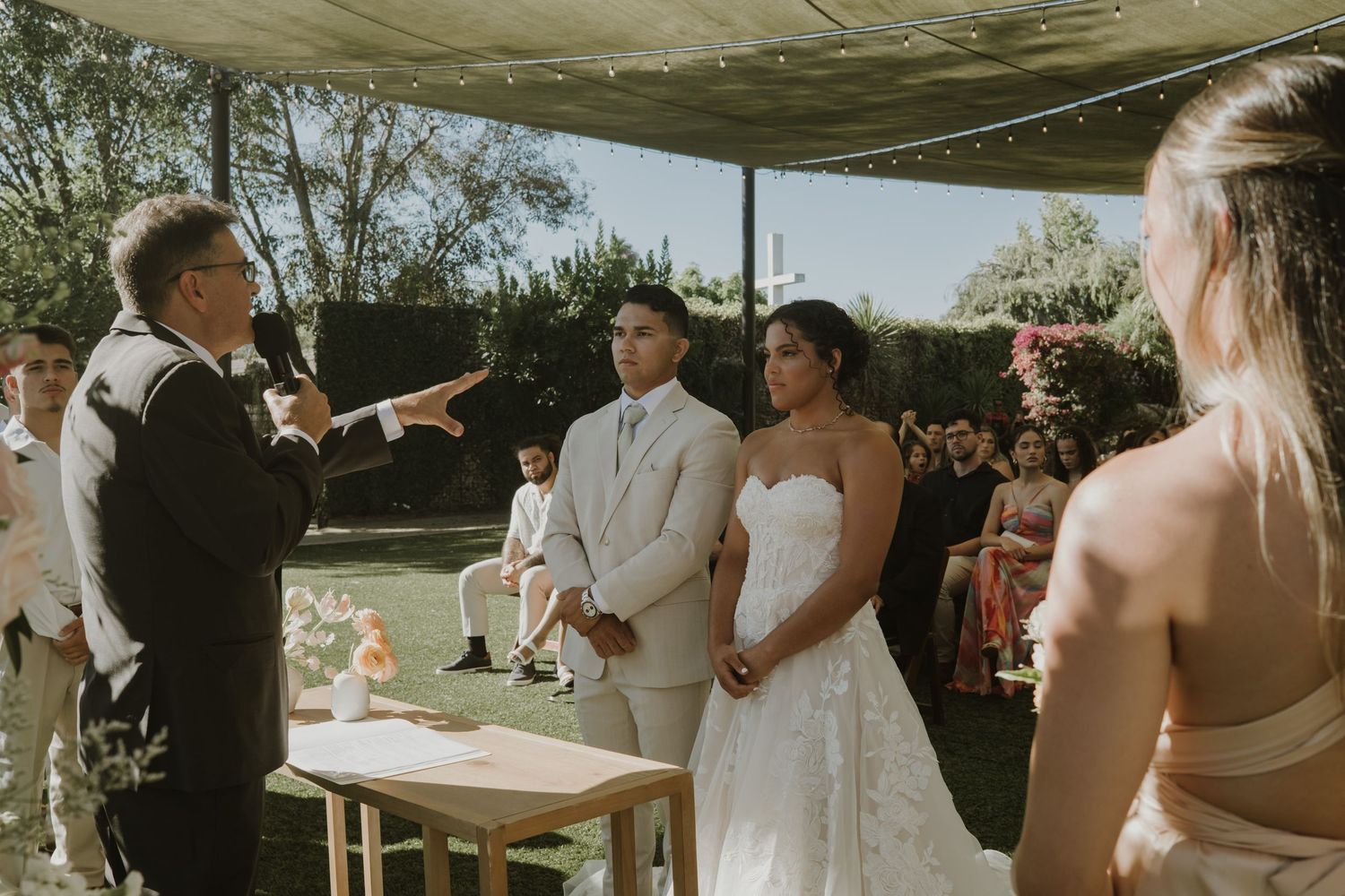 Outdoor wedding ceremony with bride and groom standing before officiant in a garden setting.