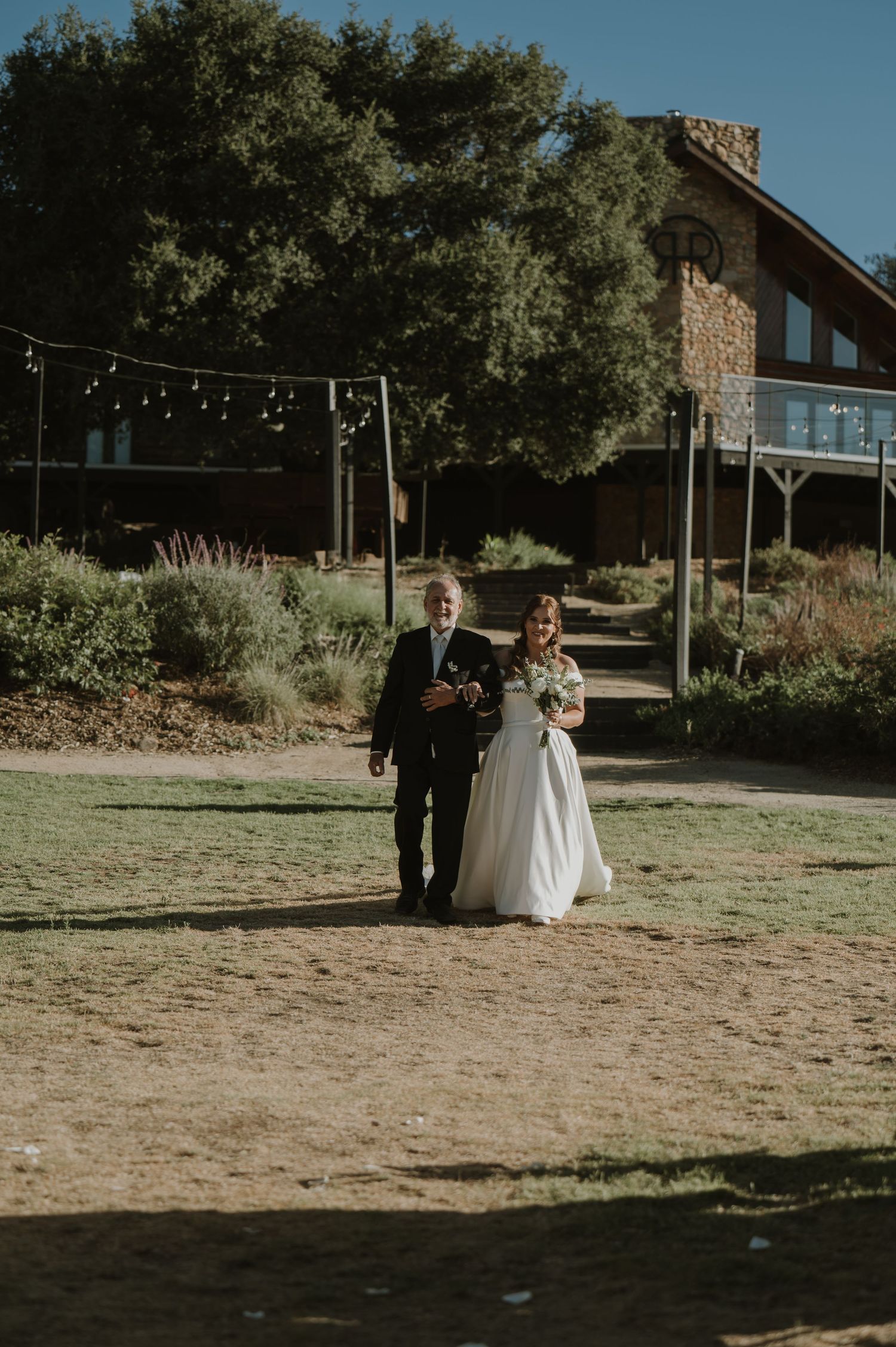Bride in white gown walks with escort toward rustic stone venue on sunny wedding day.