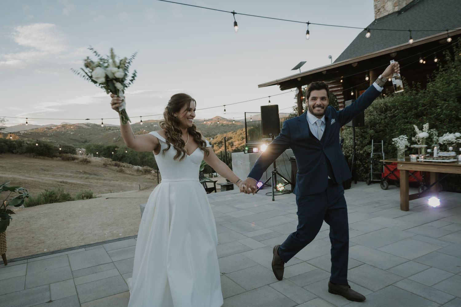 Joyful bride and groom celebrating outdoors at dusk, bride holding bouquet, groom raising bottle, rustic venue backdrop.