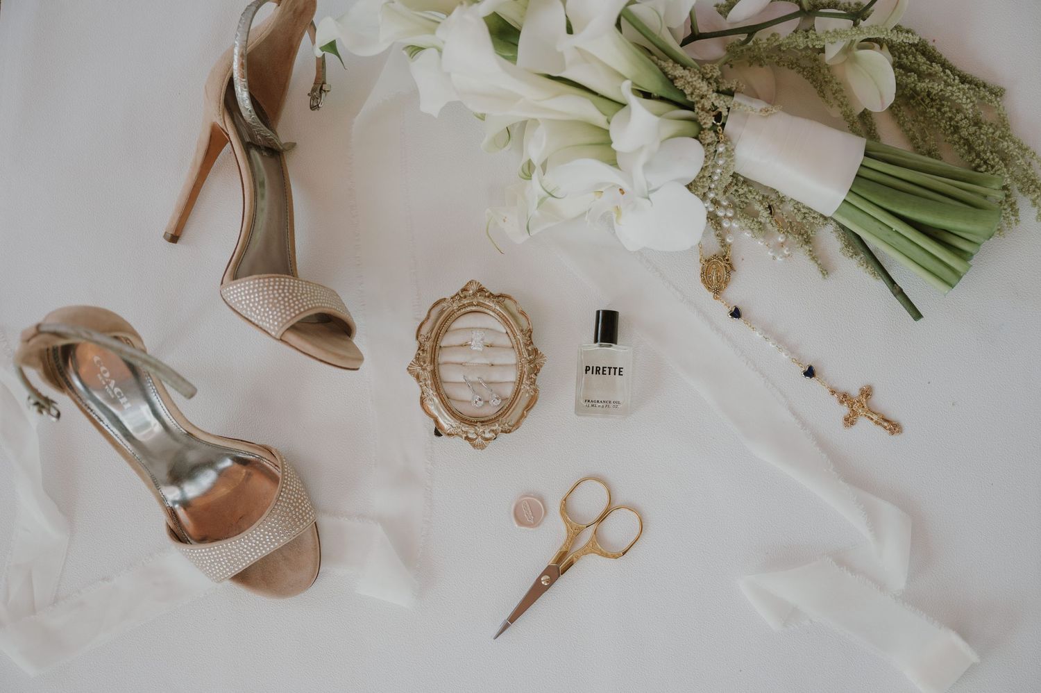 Elegant bridal flat lay with nude heels, white floral bouquet, gold perfume, scissors, and ribbon on white background.