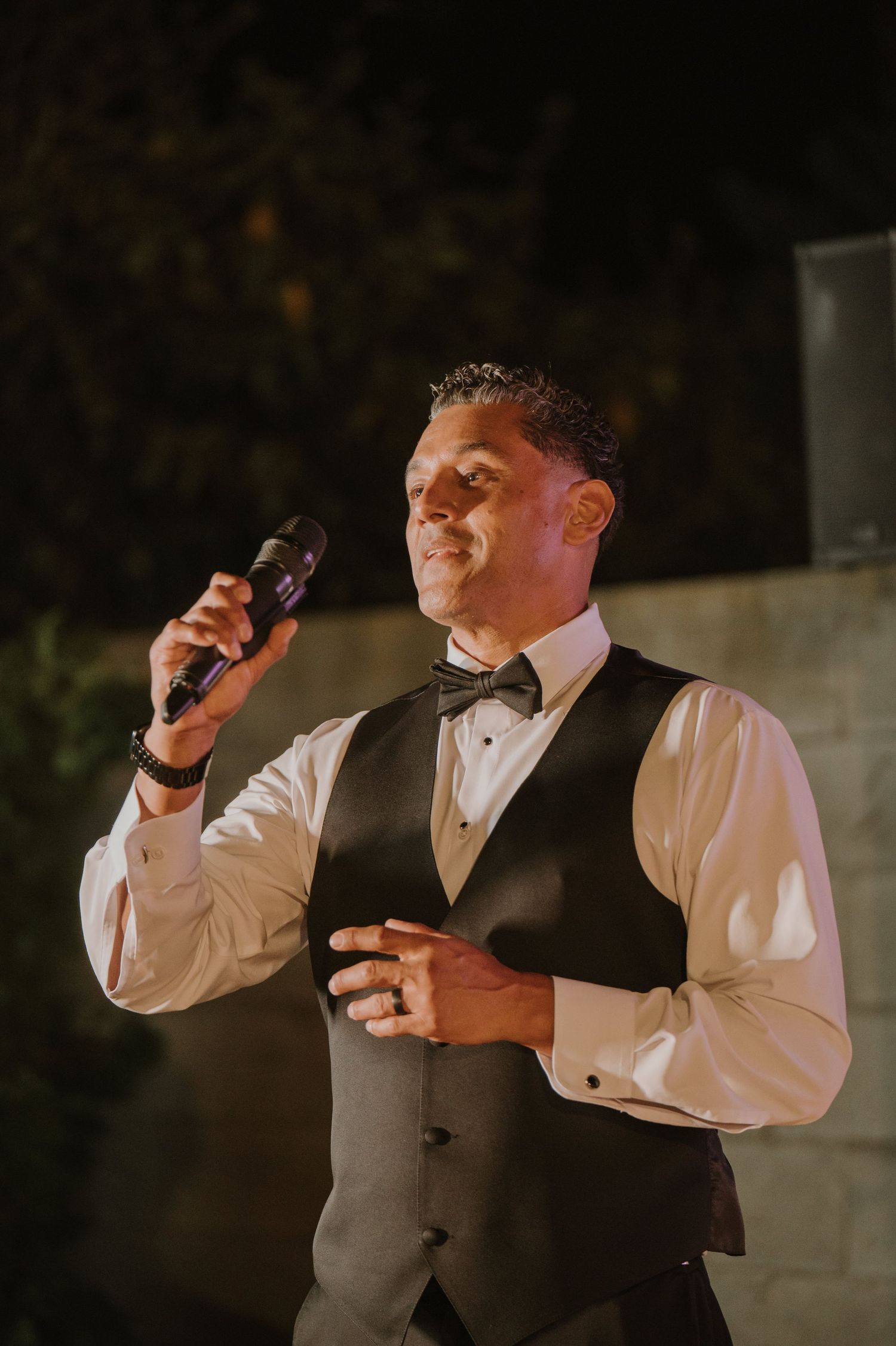 A man in a black vest and bow tie speaks into a microphone at an outdoor wedding reception.