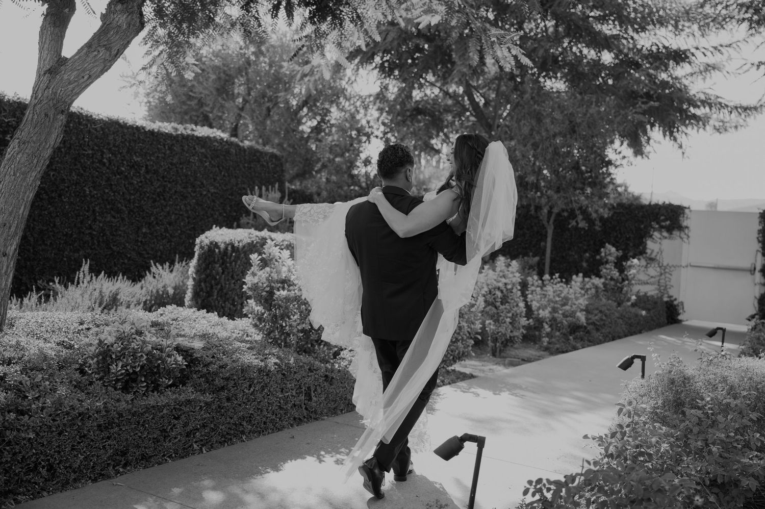 Black and white photo of groom lifting bride in wedding dress outdoors among lush greenery.