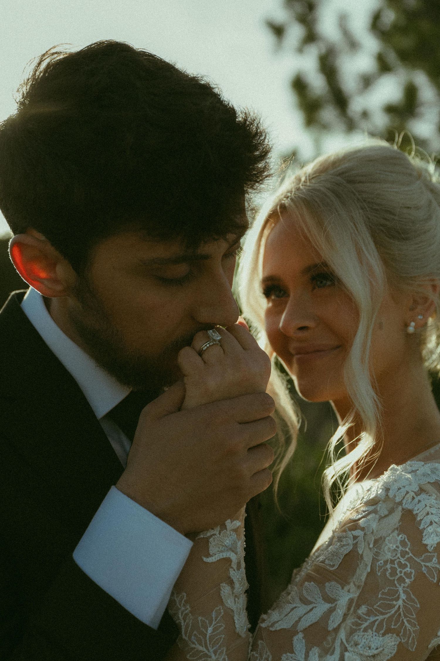 Groom in dark suit kisses bride's hand as she smiles, wearing lace wedding dress and diamond ring in golden sunlight.