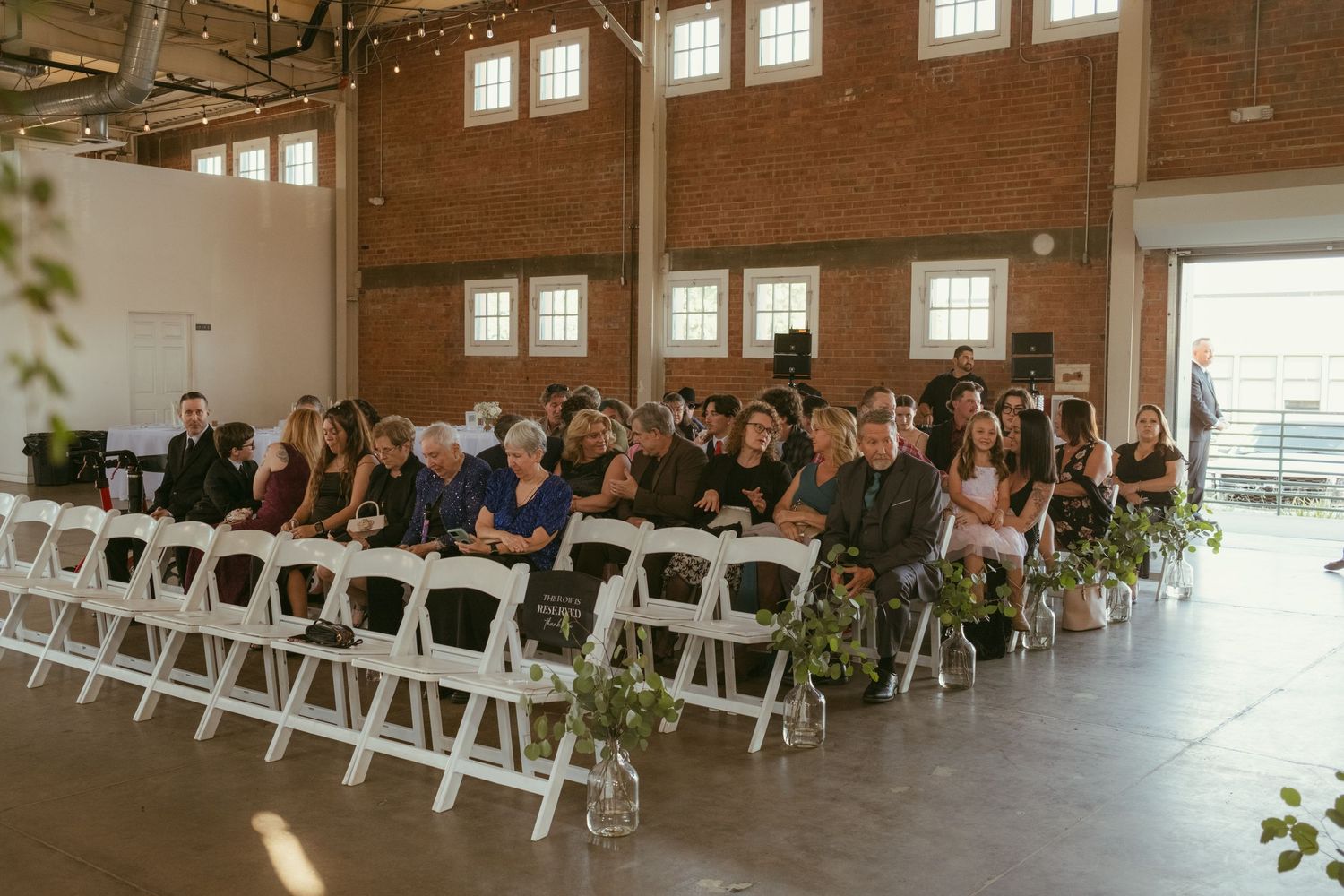 Wedding guests seated in white chairs inside rustic brick venue awaiting ceremony to begin.