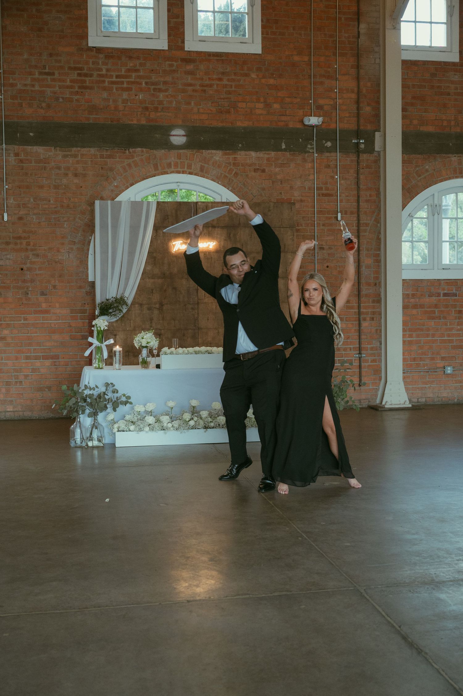 A couple in black formalwear performs an elegant dance routine in a rustic brick-walled wedding venue.