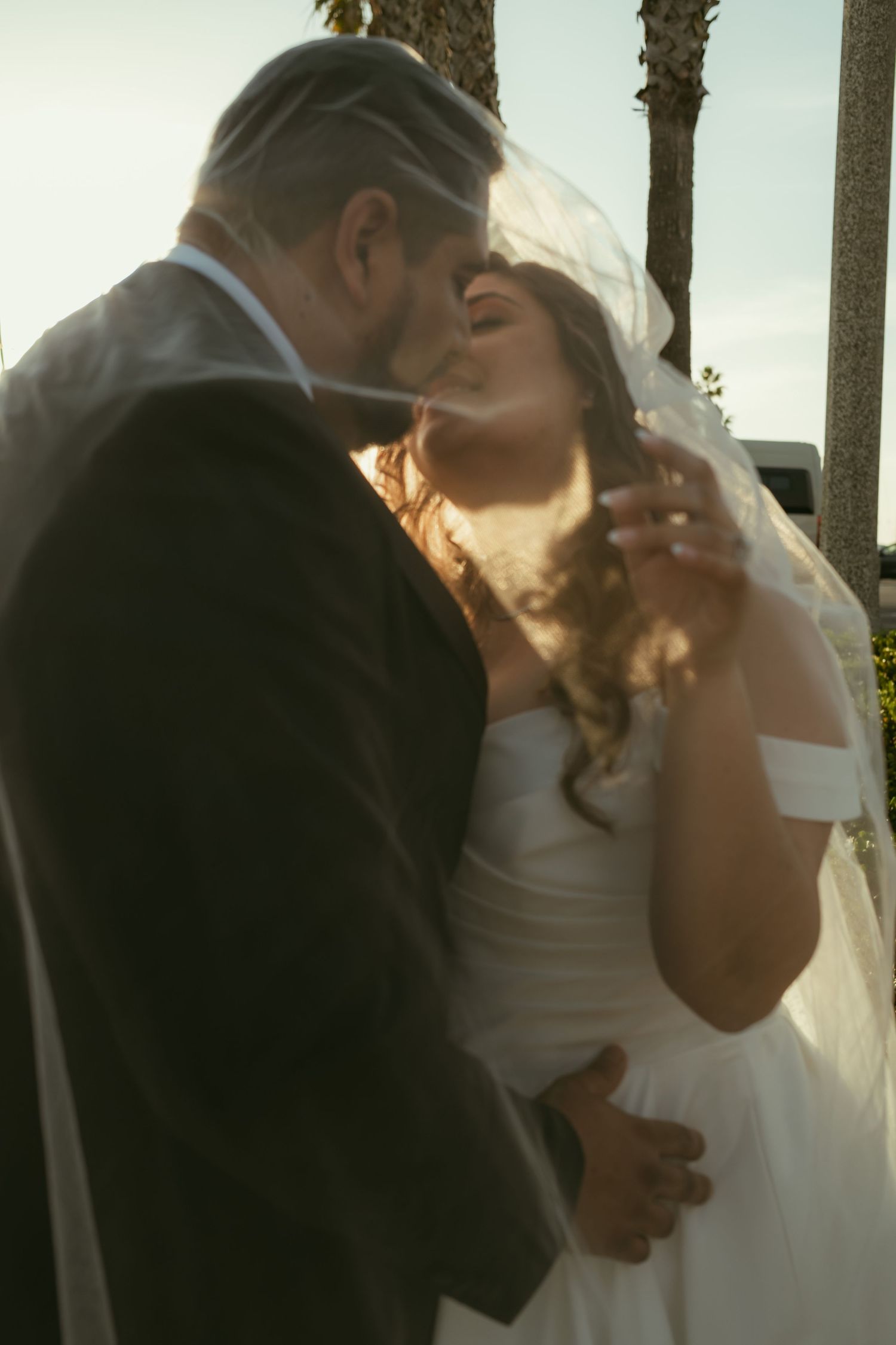 Bride and groom sharing a romantic kiss outdoors, bride in white dress and veil, groom in dark suit, golden sunlight.