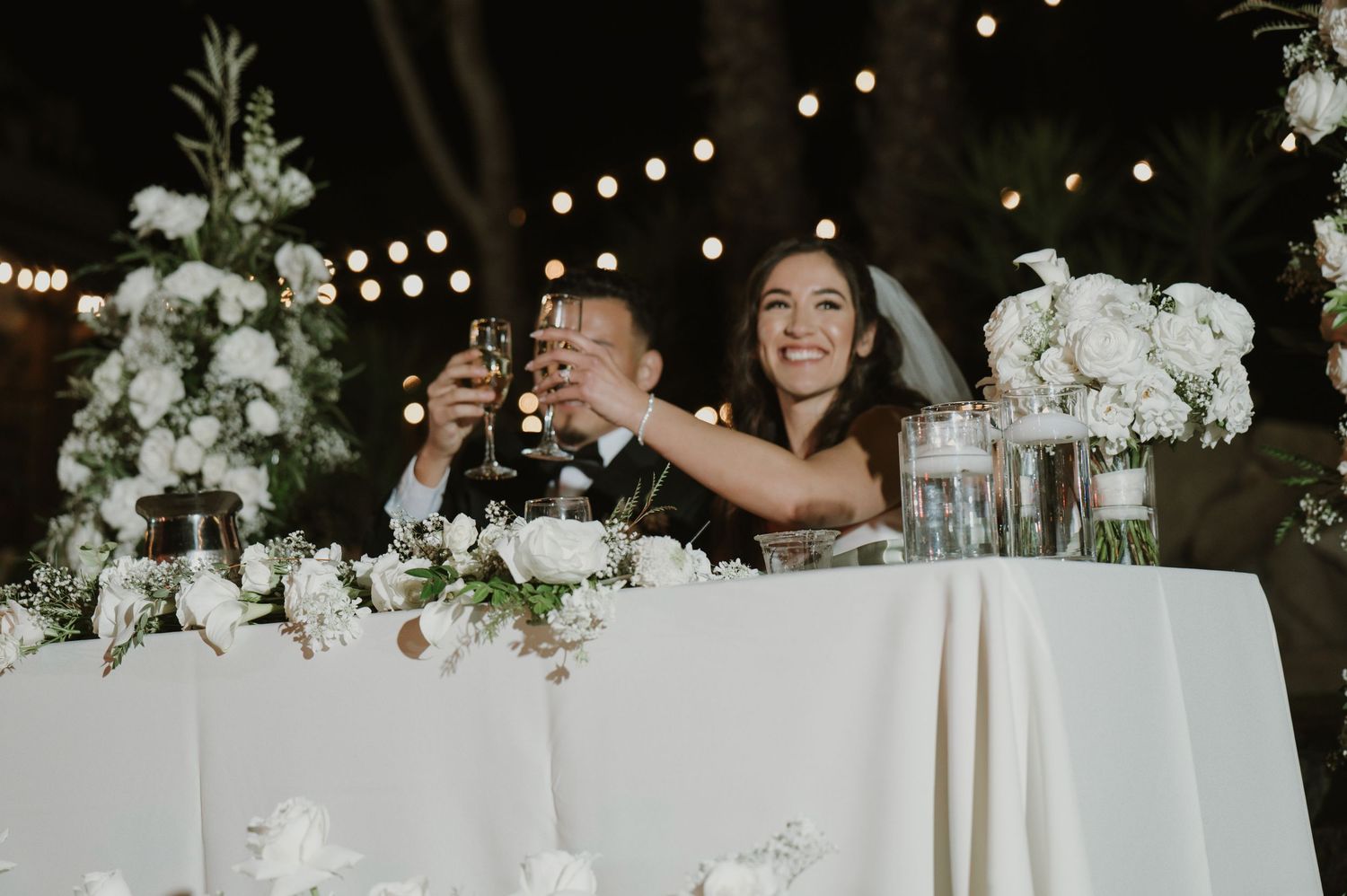 Wedding reception table decorated with white floral centerpieces and string lights overhead.