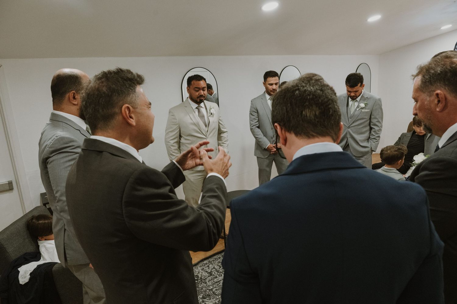 Groomsmen in gray suits gather around the groom in a bright room before the wedding ceremony.