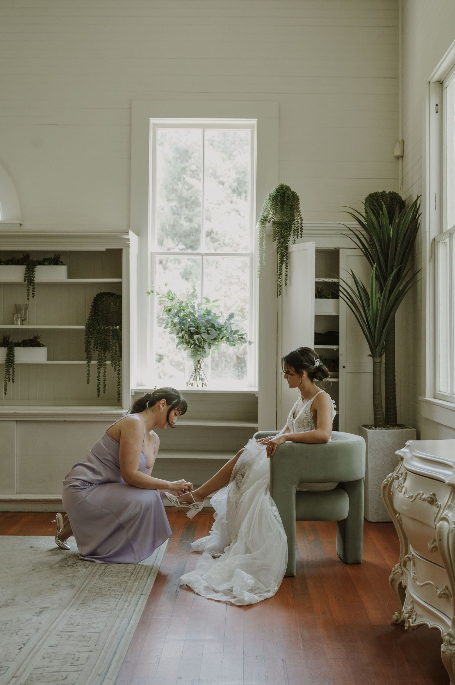Bridesmaid in lavender dress helping bride with her wedding gown train while seated in a chair.