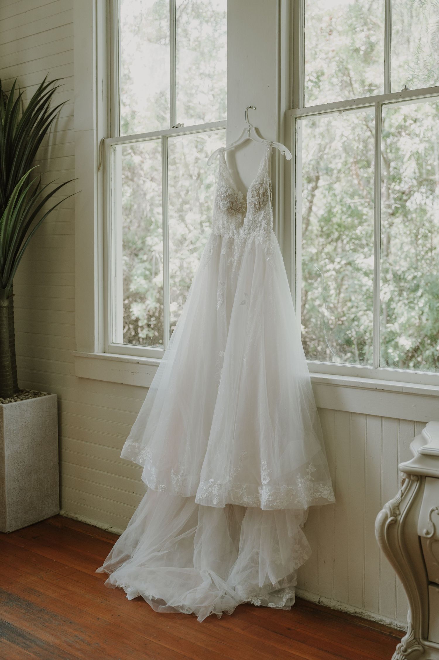 A flowing white bridal gown with lace bodice and tiered tulle skirt hanging by a sunlit window.