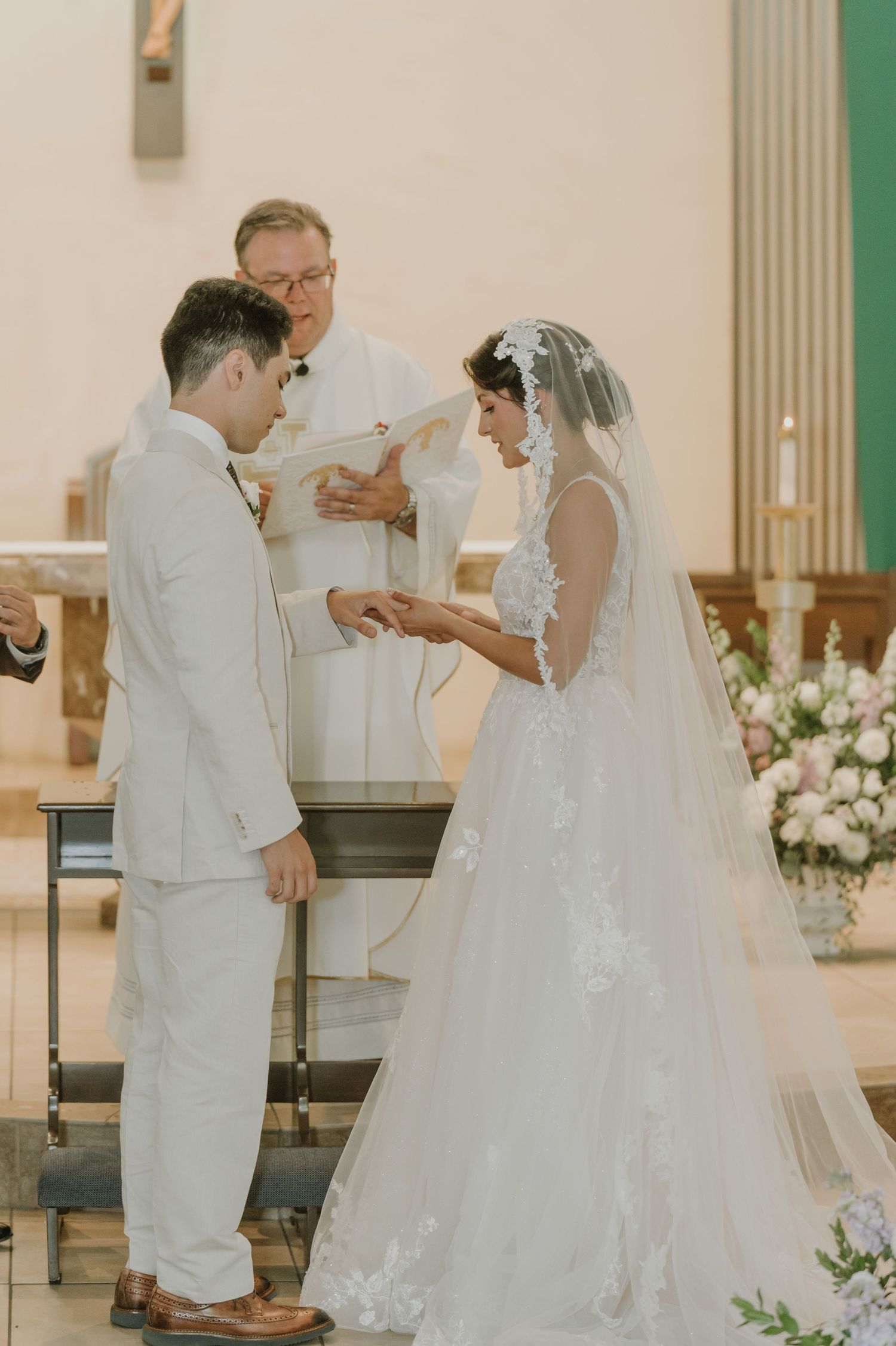 Bride in lace gown and veil exchanges rings with groom in white suit before officiant at altar.