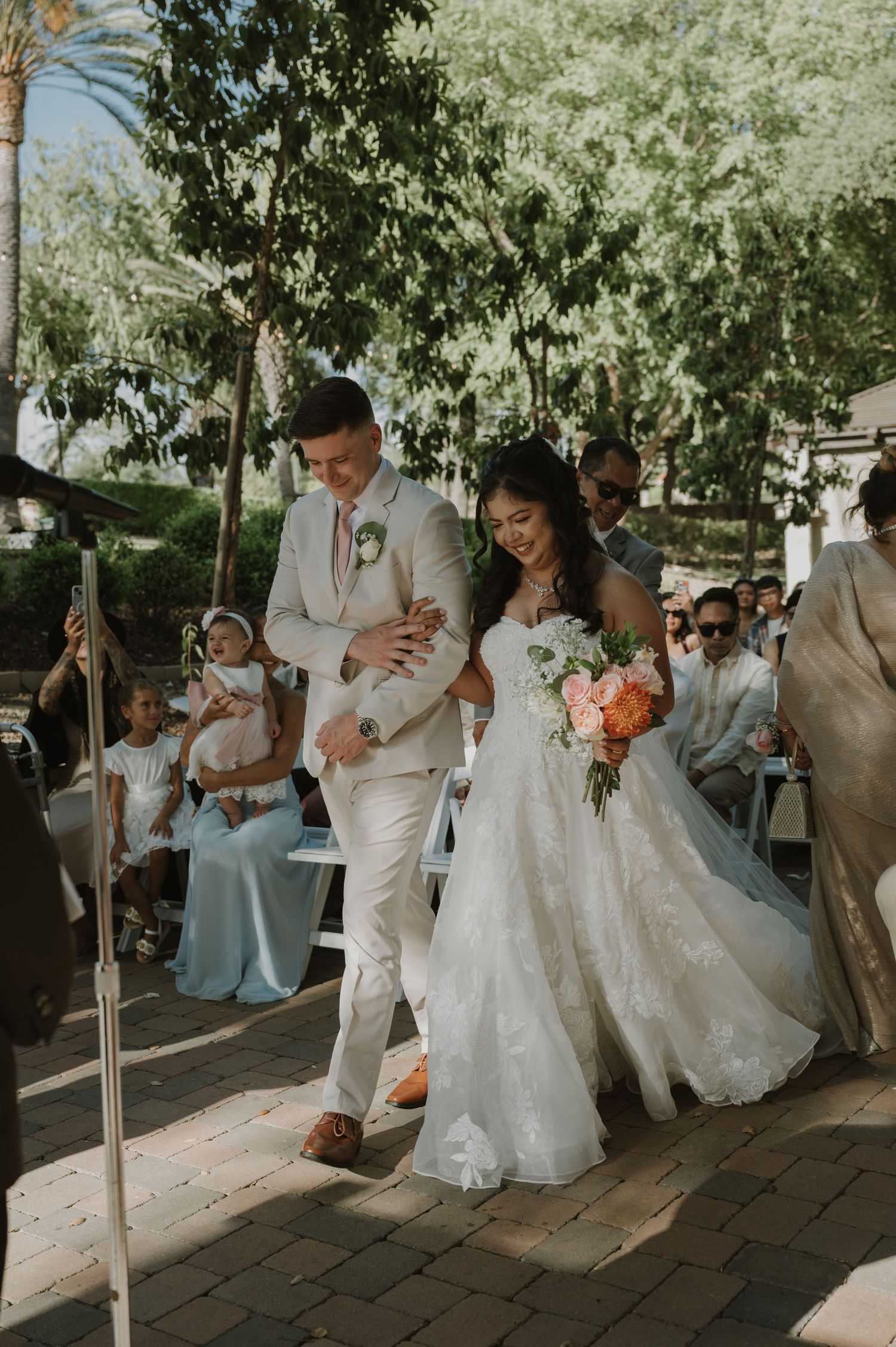 Bride and groom walk down the aisle at an outdoor wedding ceremony, bride holding a colorful bouquet.