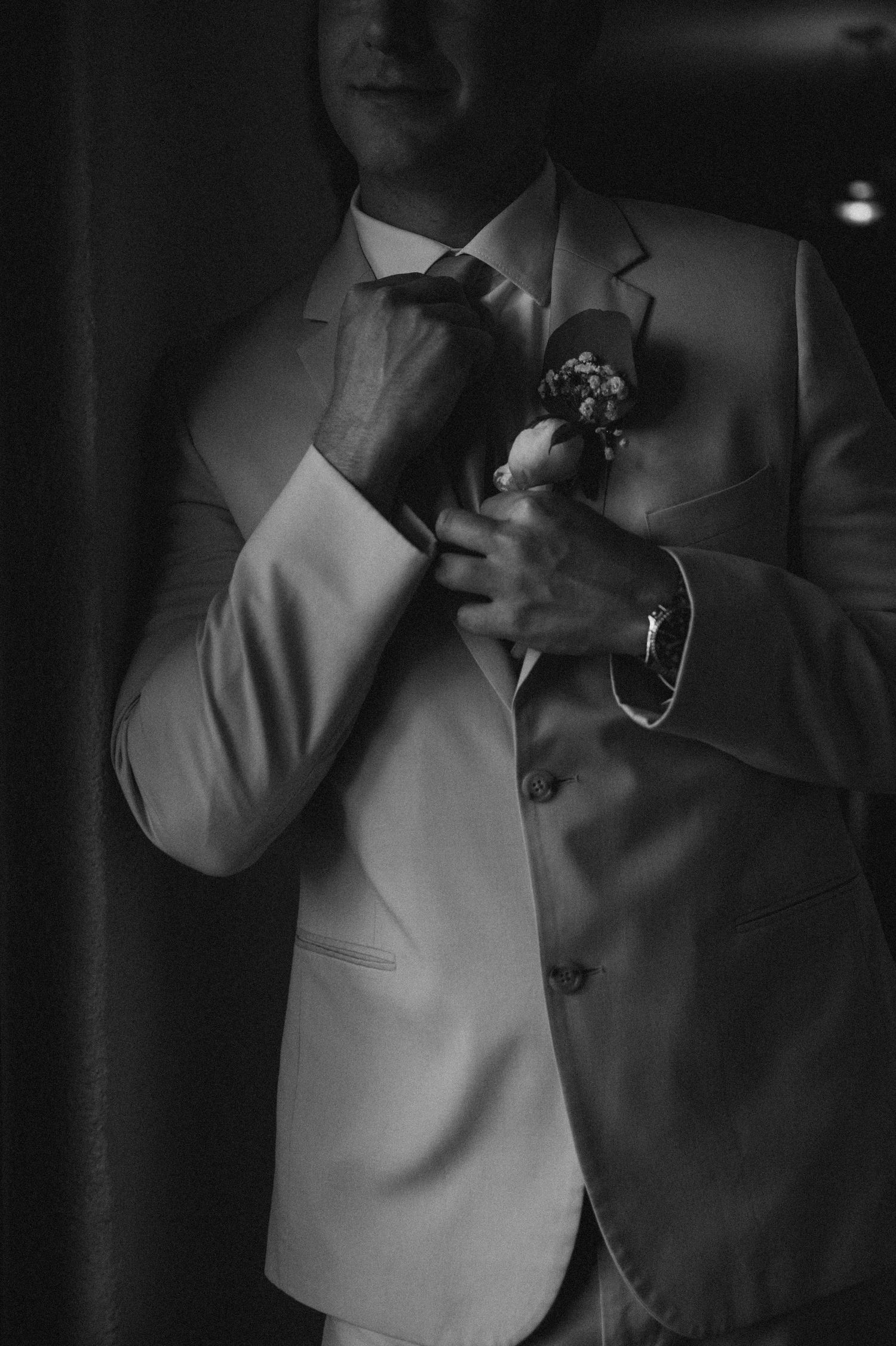 Black and white photo of a groom in a suit adjusting his boutonniere before his wedding ceremony.