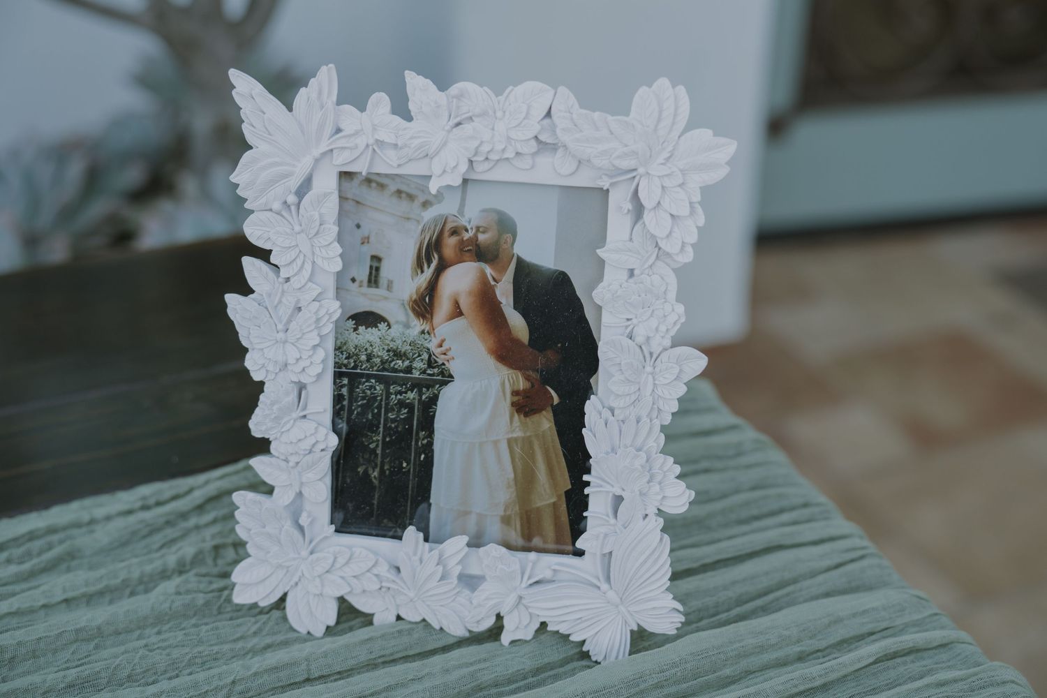 White ornate leaf-decorated photo frame displaying a wedding couple kissing, placed on a teal surface.