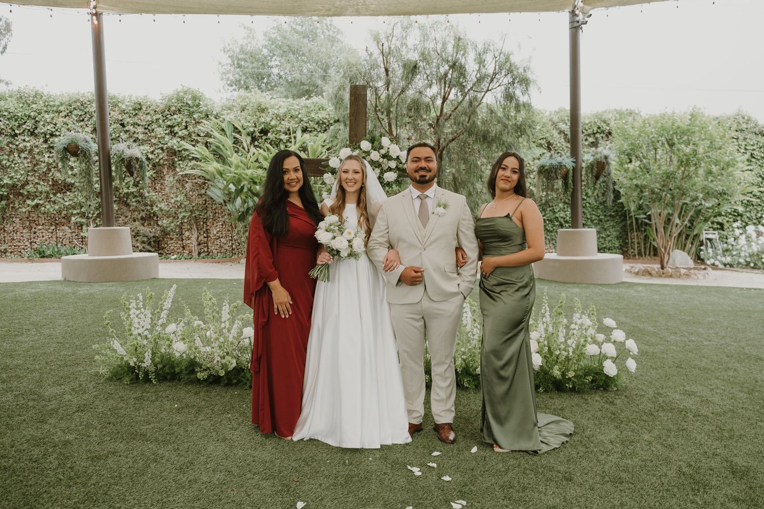 Bride and groom pose with two bridesmaids in green and red dresses at an outdoor garden wedding ceremony.