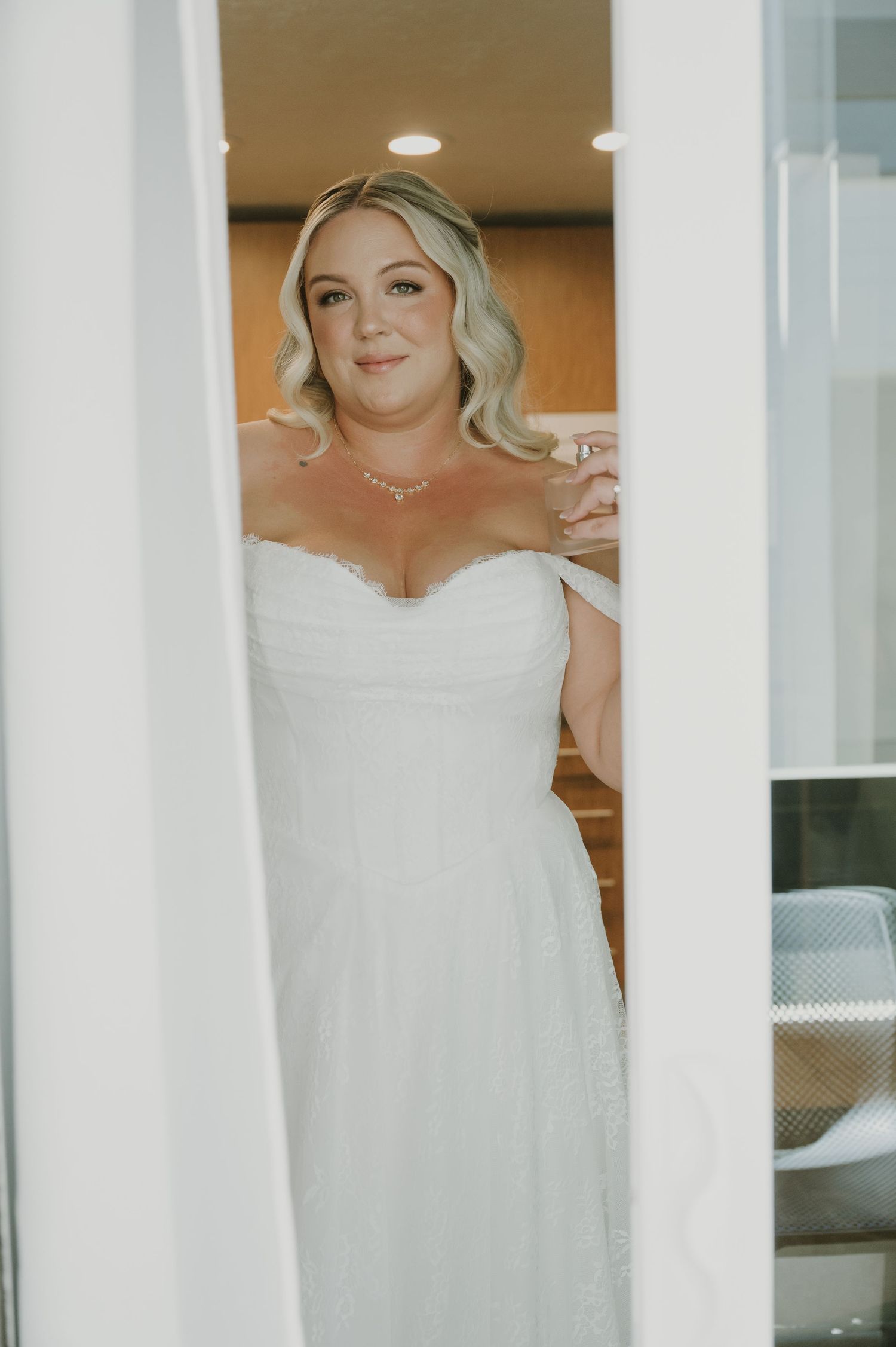 Bride in white off-shoulder wedding gown smiling through doorway, blonde hair, elegant necklace, soft natural lighting.