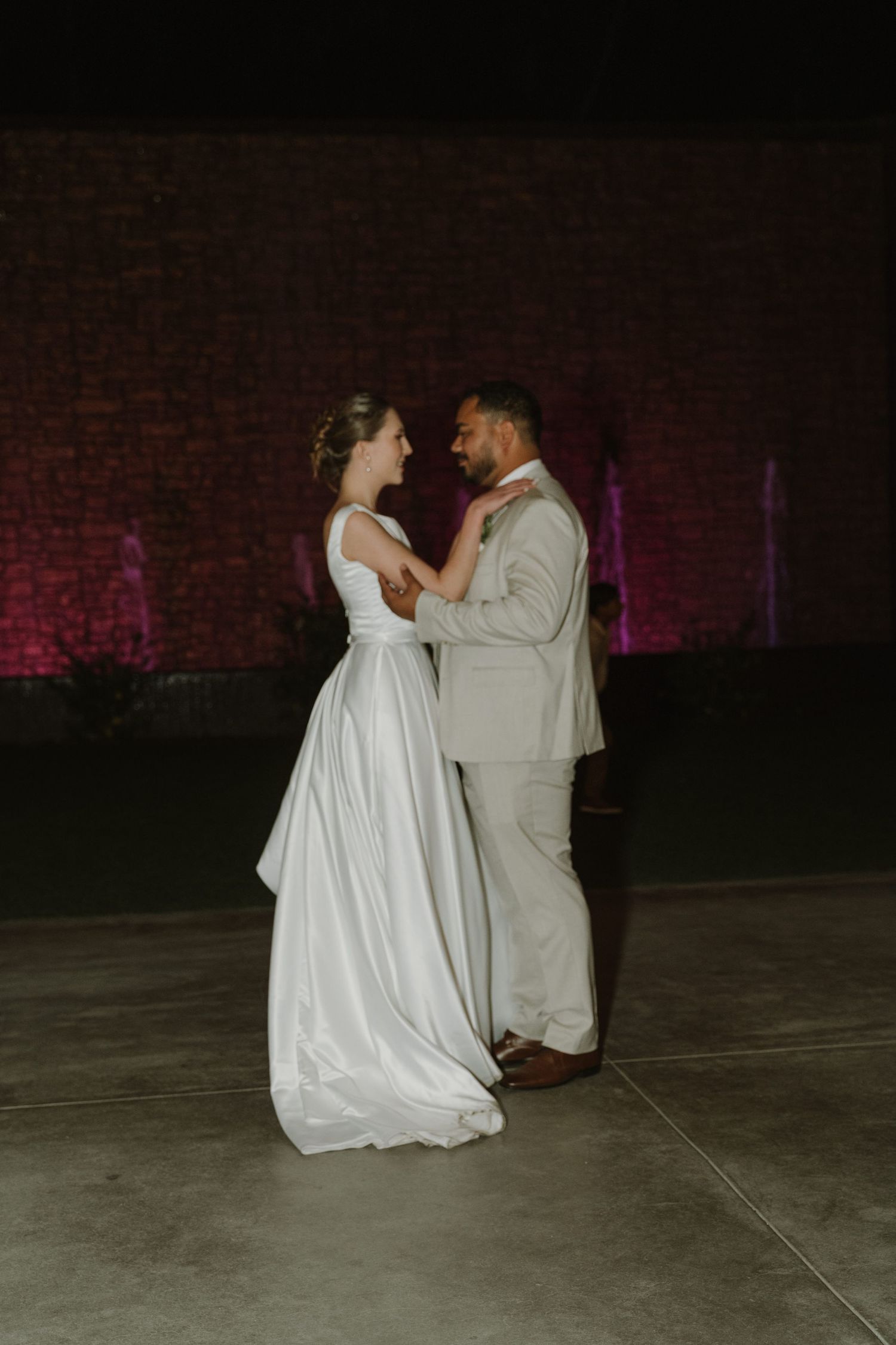 Bride in white satin gown and groom in beige suit share their first dance at an outdoor evening wedding reception.