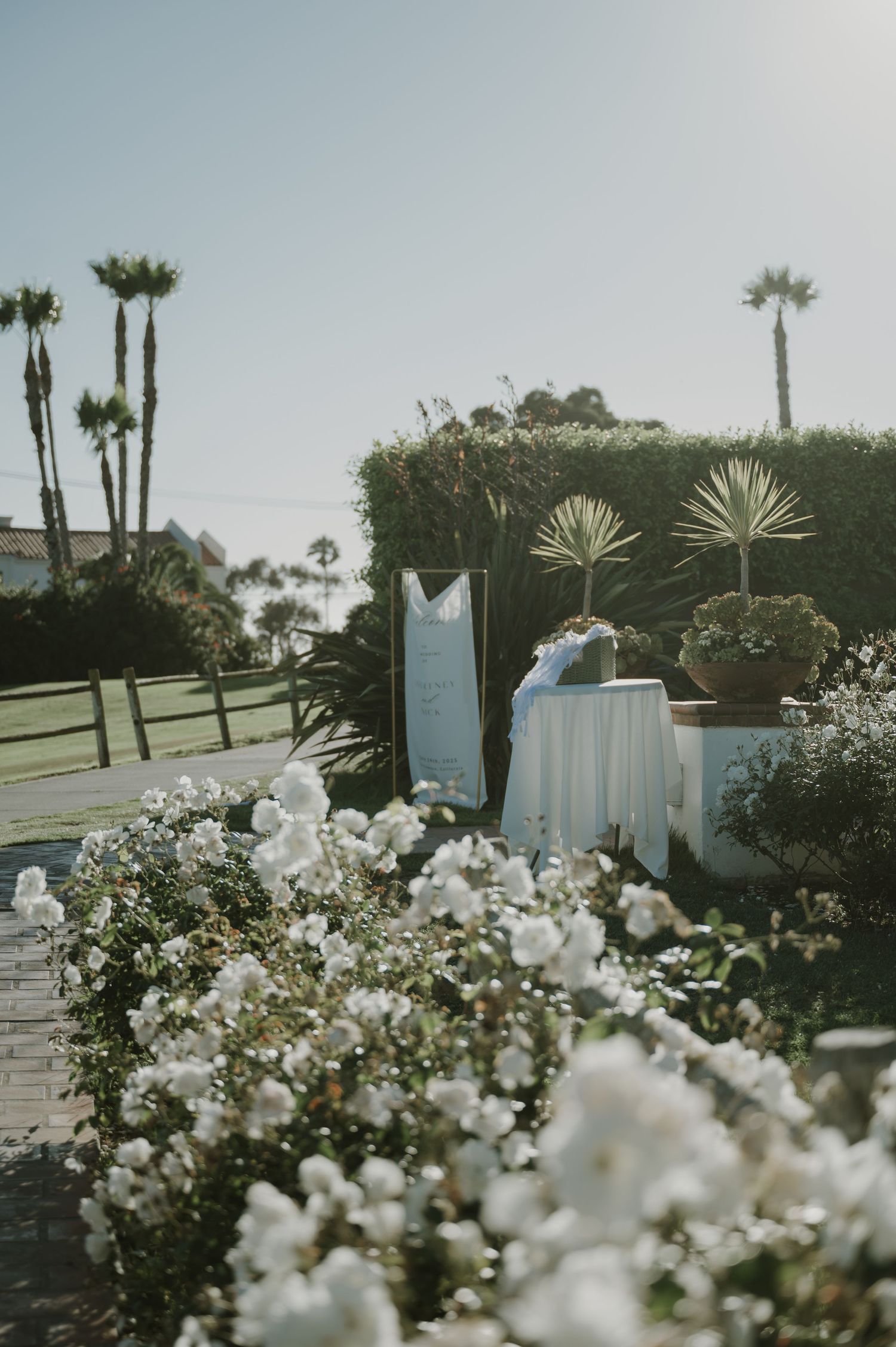 Outdoor wedding setup with white roses, palm trees, and elegant table decor under a clear sky.