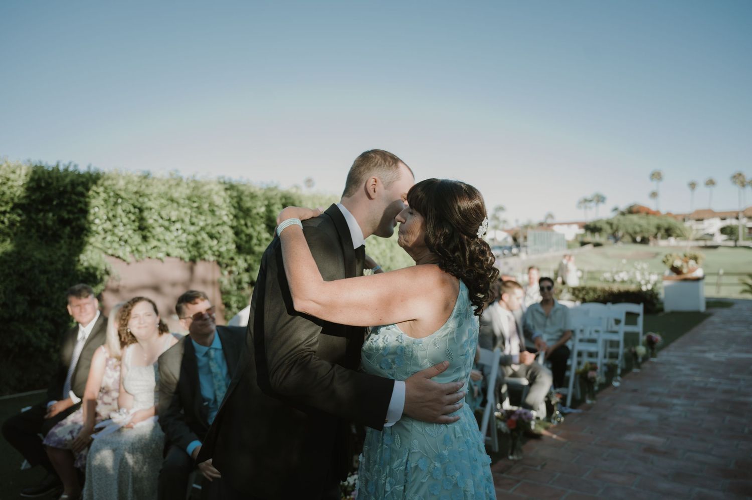 Bride and groom share first kiss at outdoor wedding ceremony with guests seated on sunny day.