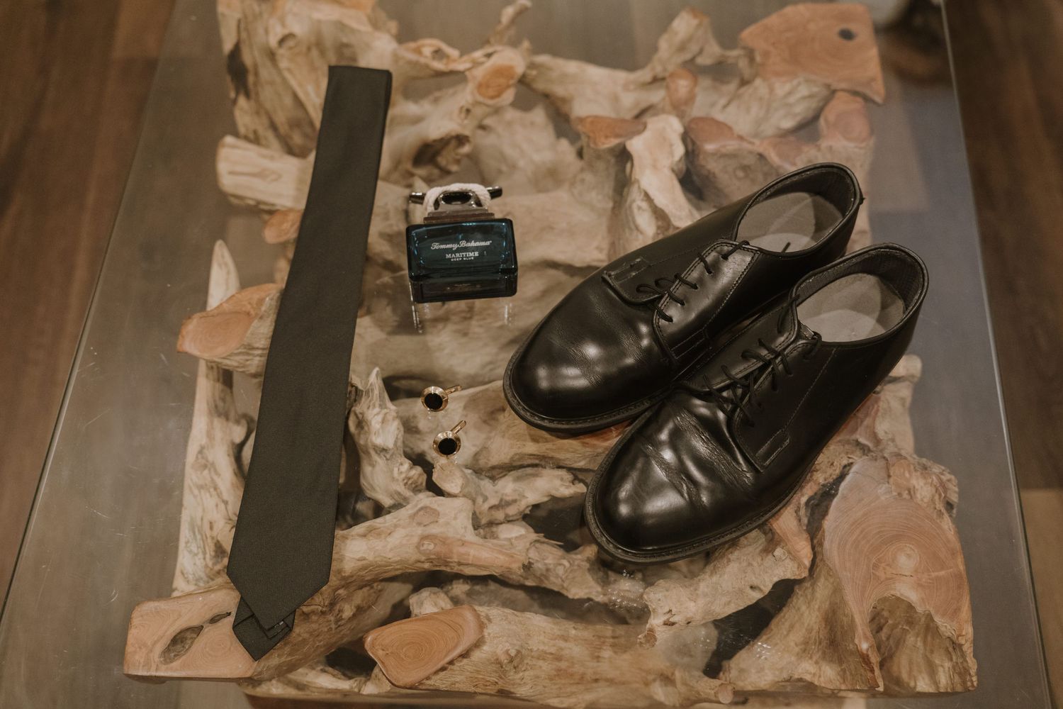 Black leather dress shoes, olive tie, and cologne displayed on driftwood for a classic mens formal accessories flat lay.