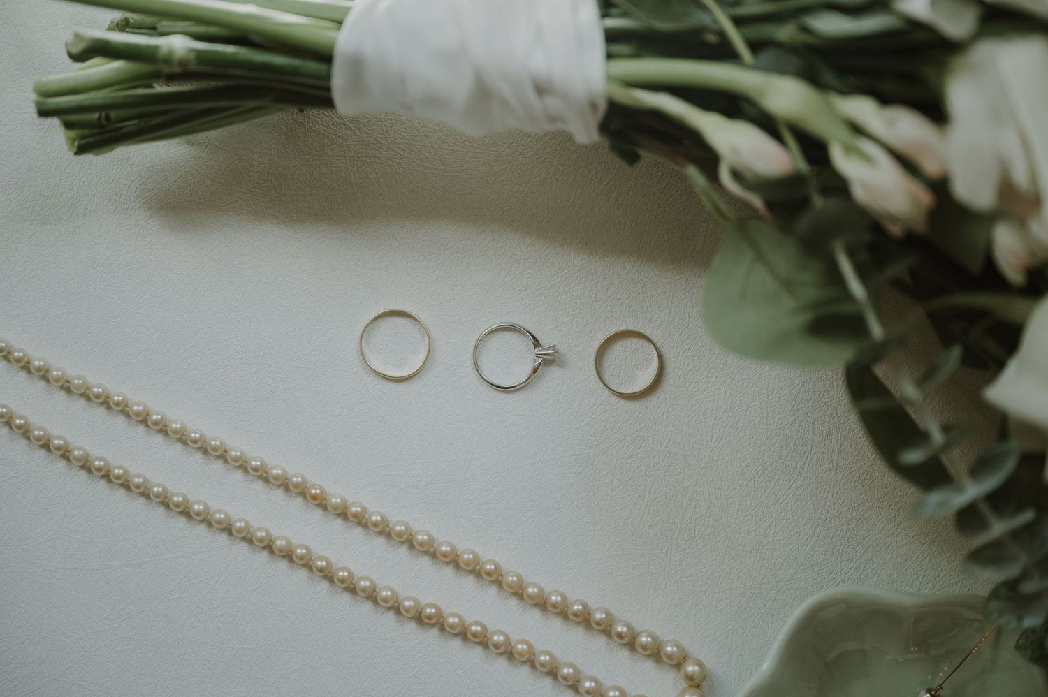 Three gold wedding rings and a pearl necklace arranged near a bridal bouquet of white flowers on a light surface.