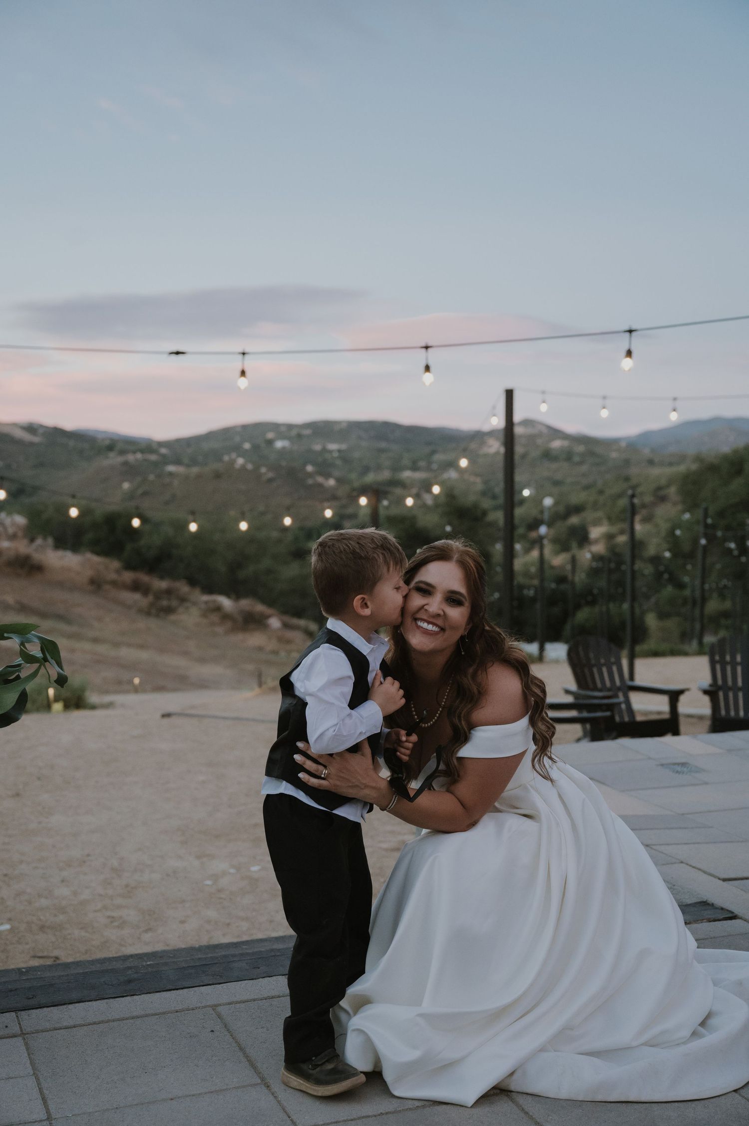Smiling bride in white gown hugs a young boy at an outdoor wedding with string lights and hills.
