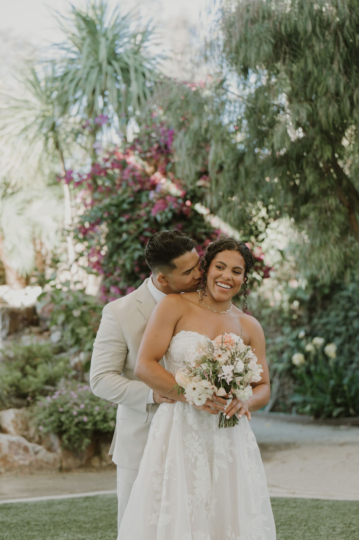 Smiling bride holding bouquet embraced by groom in tan suit, posing before lush garden with colorful bougainvillea.