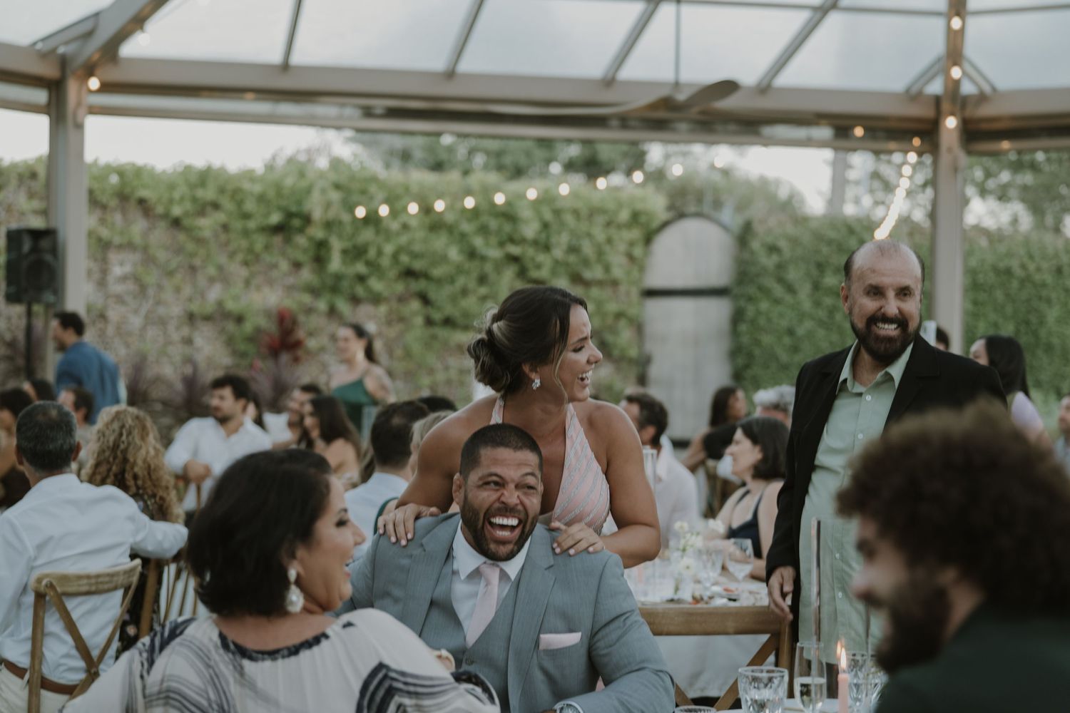 Joyful wedding reception guests laughing and celebrating together under a pergola with string lights and lush greenery.