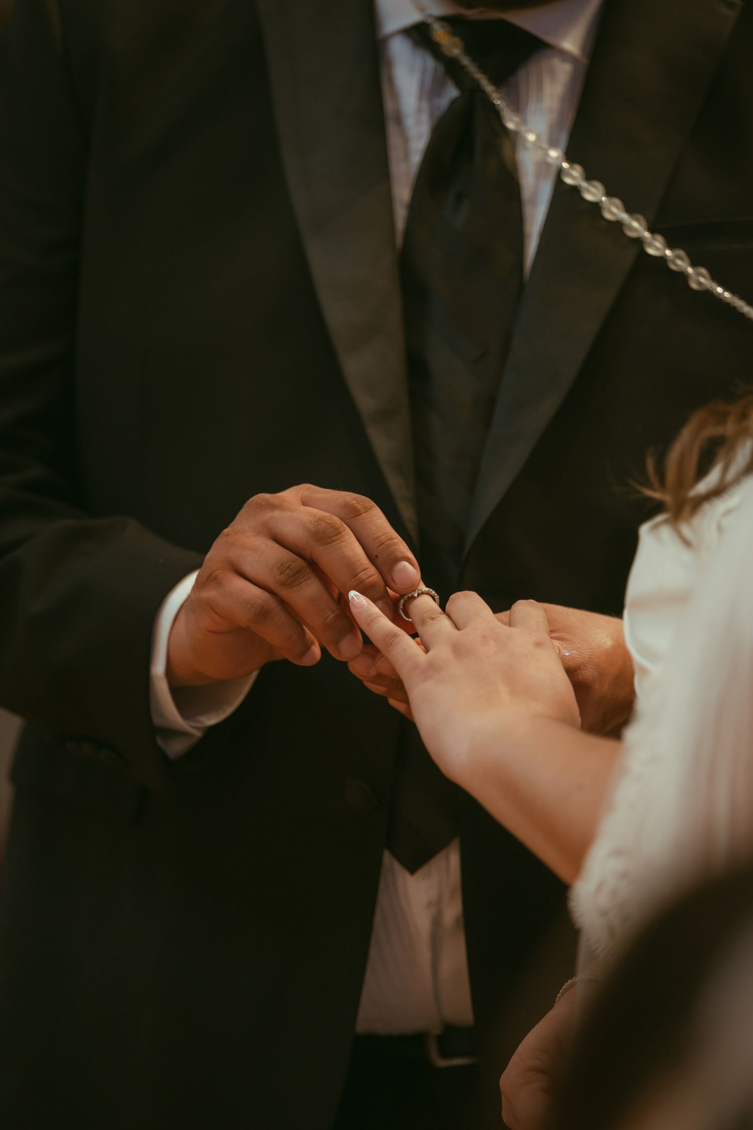 Groom placing a wedding ring on bride's finger during ring exchange ceremony, wearing black suit.