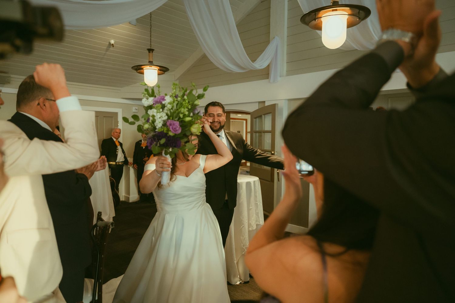 Bride and groom dance at wedding reception as guests celebrate and photograph the joyful moment.
