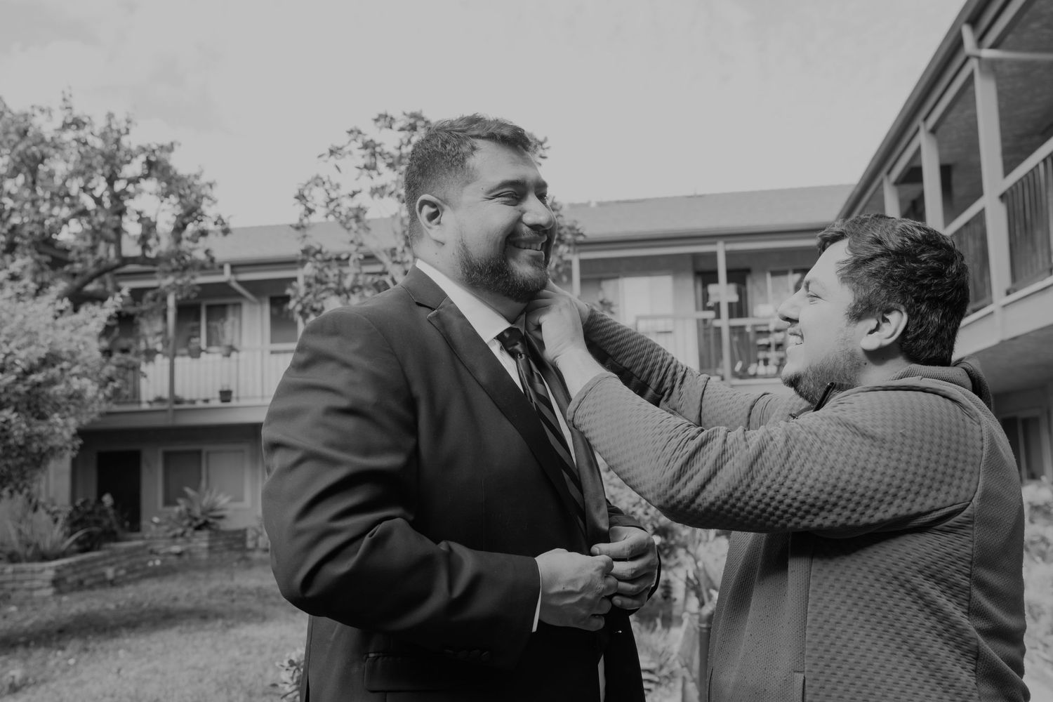 A black and white photo of two men sharing a joyful moment outdoors, one adjusting the other's tie before a wedding.