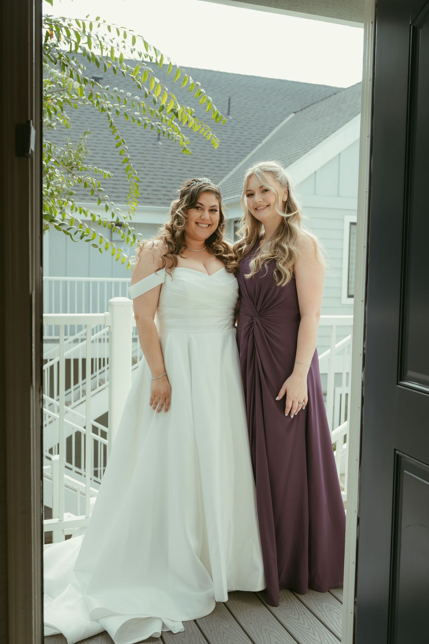 Bride in white off-shoulder gown poses with bridesmaid in purple dress on wooden deck at outdoor wedding venue.