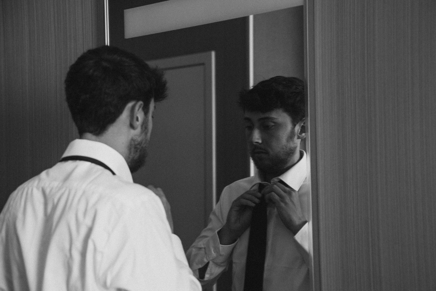 Groom adjusting tie in mirror reflection wearing white dress shirt in black and white photo.