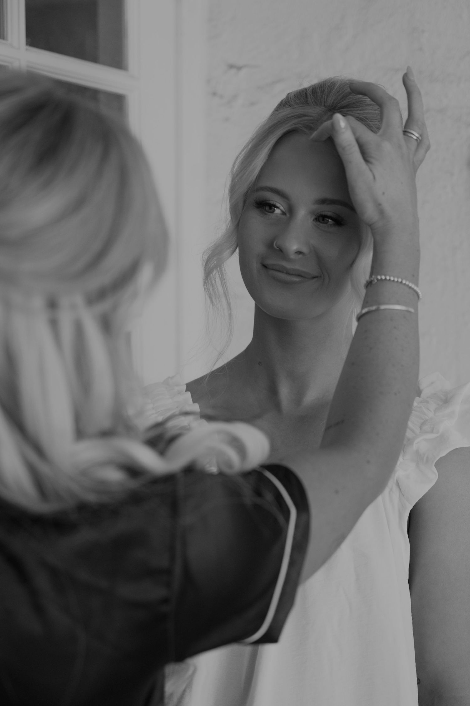 Black and white portrait of a smiling bride adjusting her hair with help from her bridesmaid.