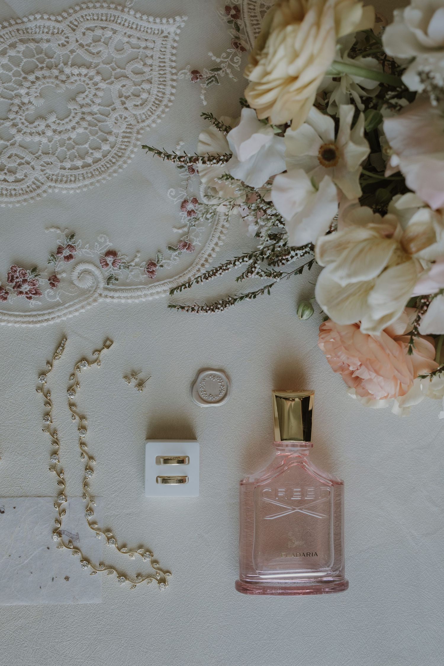 Elegant bridal flat lay featuring Creed perfume, pearl bracelet, gold ring, earrings, and fresh blush flowers on white surface.