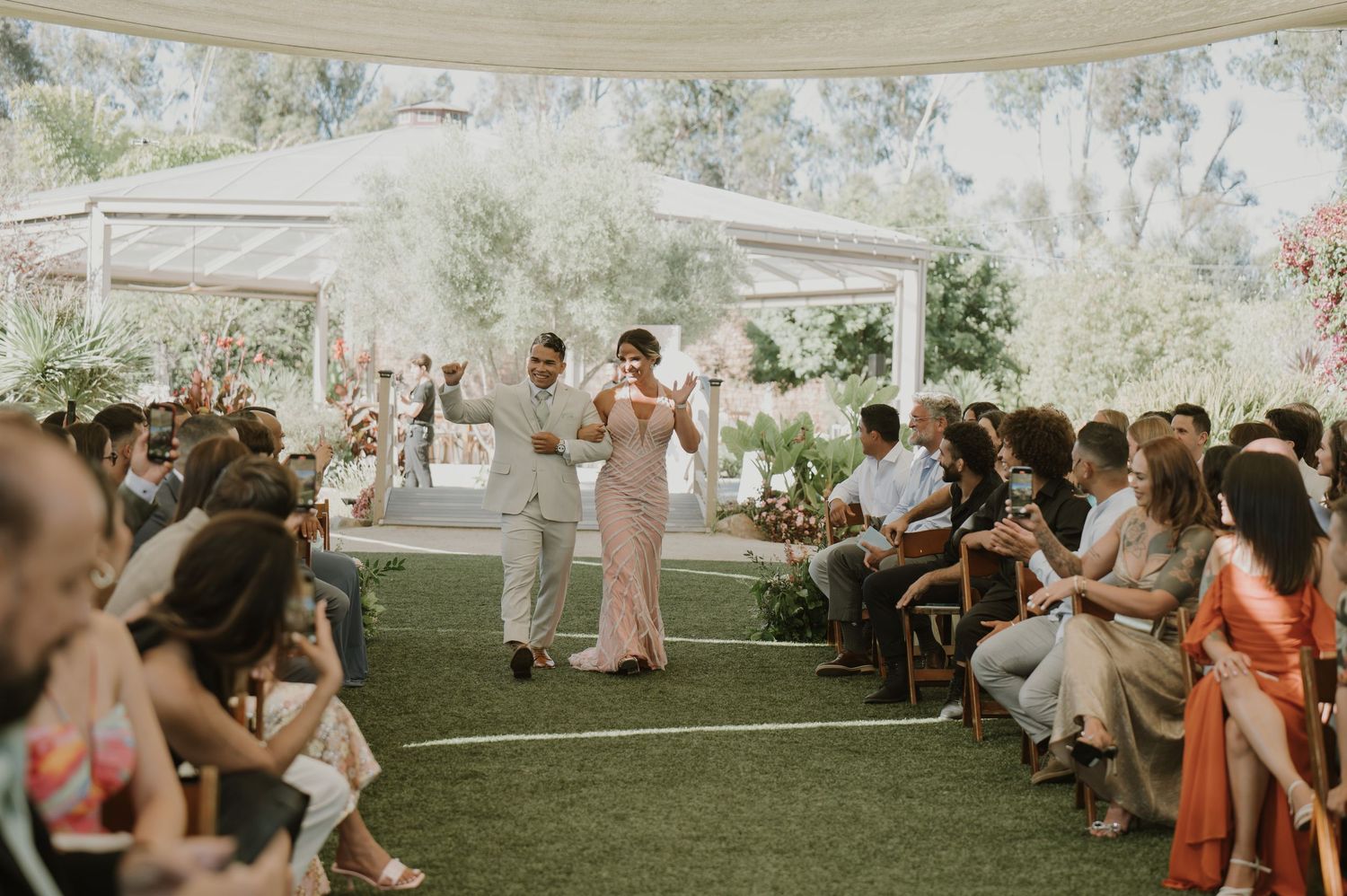 Couple walks down the aisle at an outdoor wedding ceremony under a white tent with guests seated on both sides.