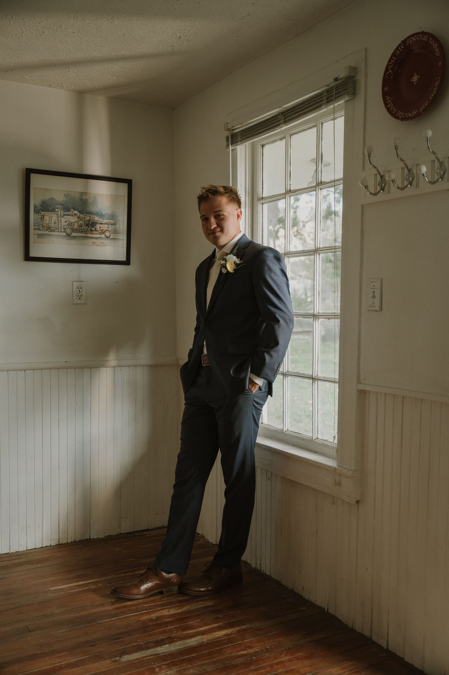A person in formal attire stands near a window in a room with white wainscoting and hardwood floors.
