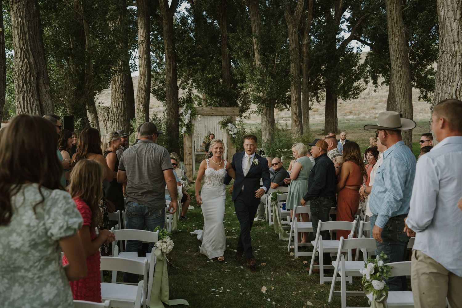 Wedding guests gather on a tree-lined lawn with white chairs set up for an outdoor ceremony.