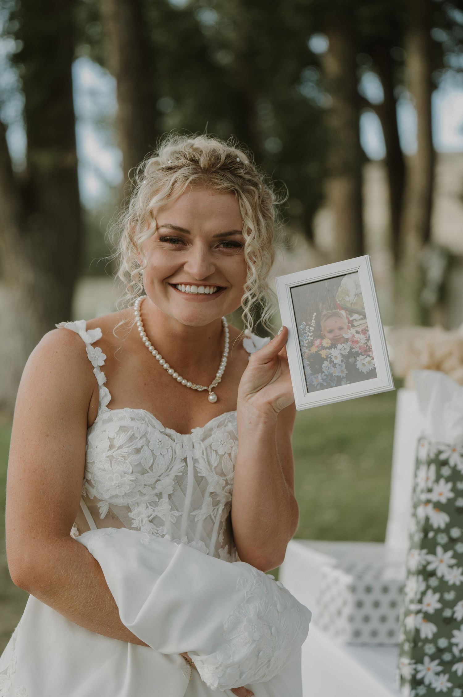 Bride in lace dress poses joyfully outdoors while holding polaroid photos during wedding photoshoot.
