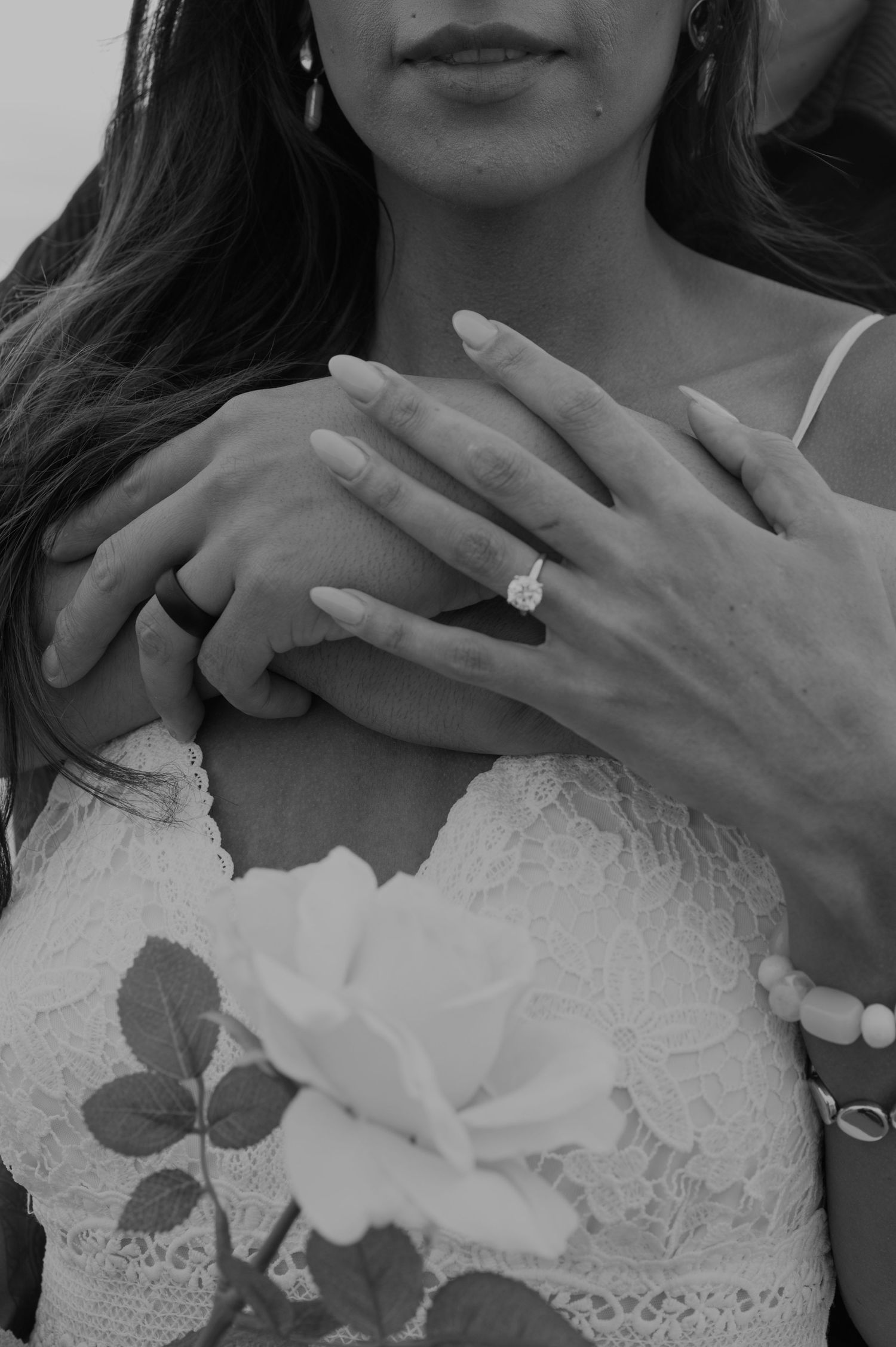 Close-up of hands with engagement ring and lace wedding dress details in black and white.