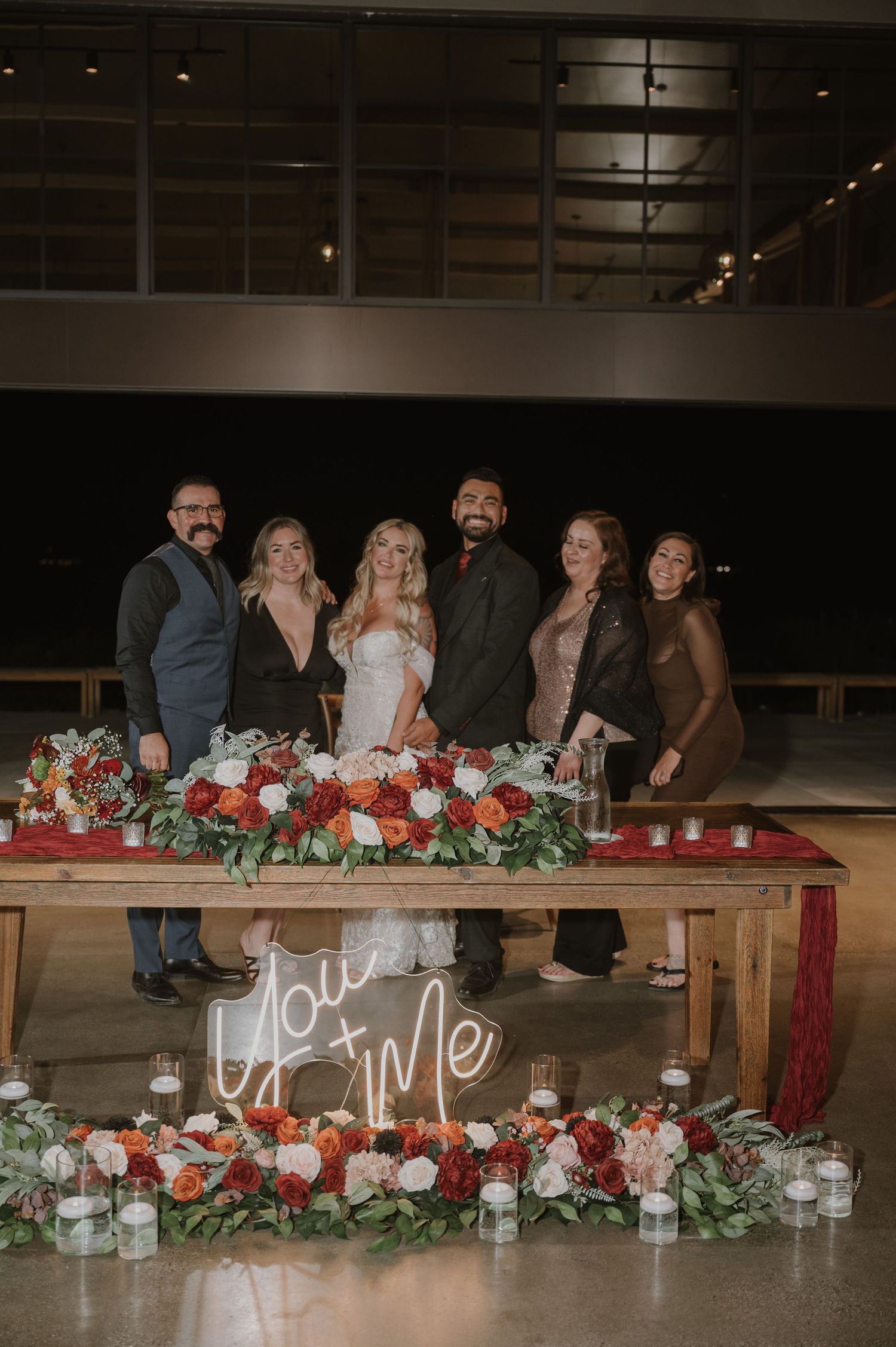 Long wooden tables adorned with red floral centerpieces and white candles in an industrial venue.