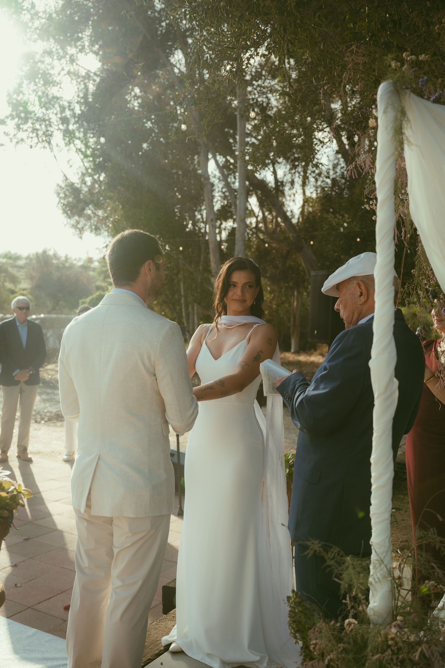 Bride and groom exchange vows outdoors at a sunlit wedding ceremony with an officiant and guests in background.