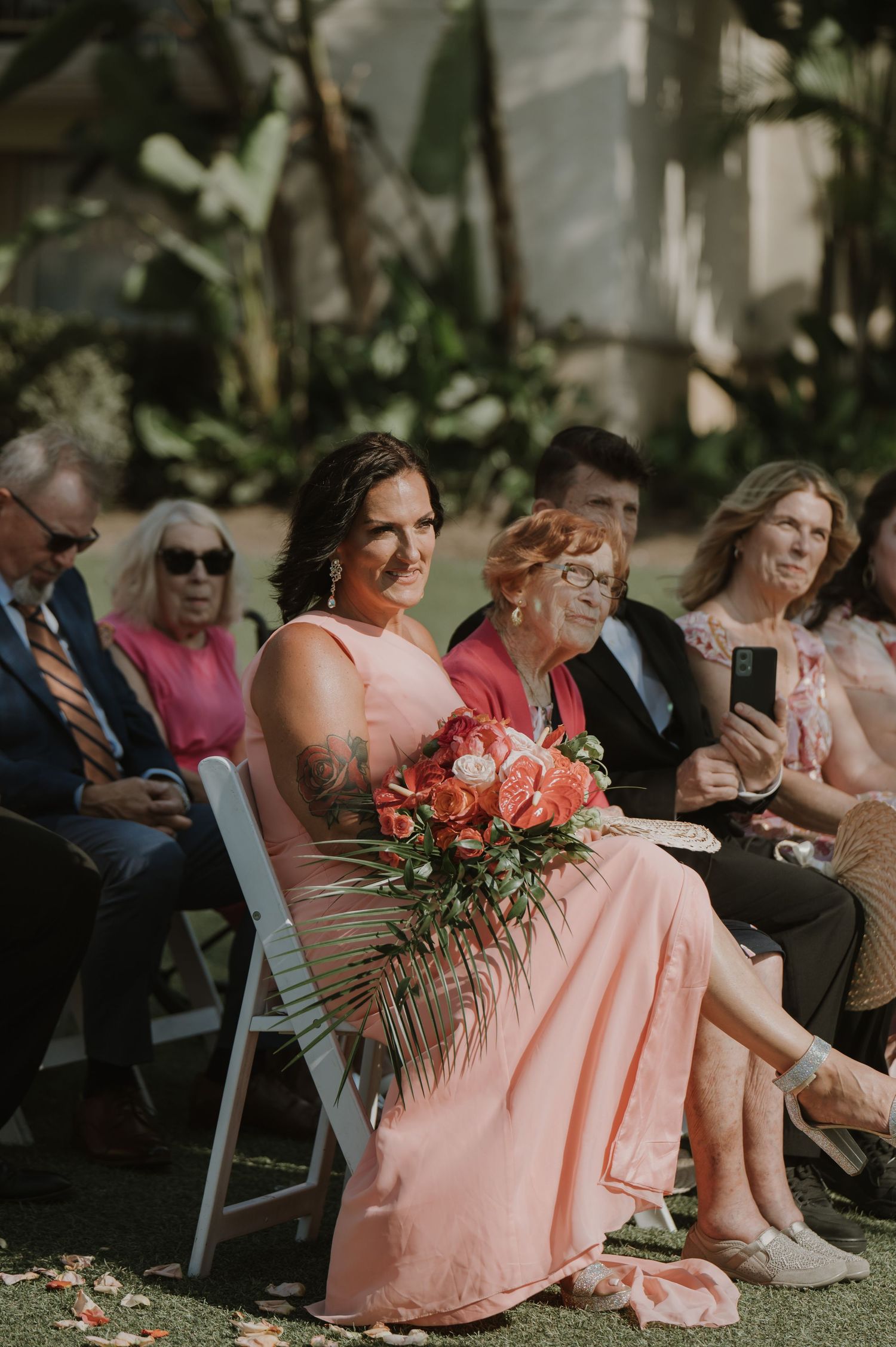Wedding guests in pink dresses sit in white chairs at an outdoor ceremony with coral floral arrangements.