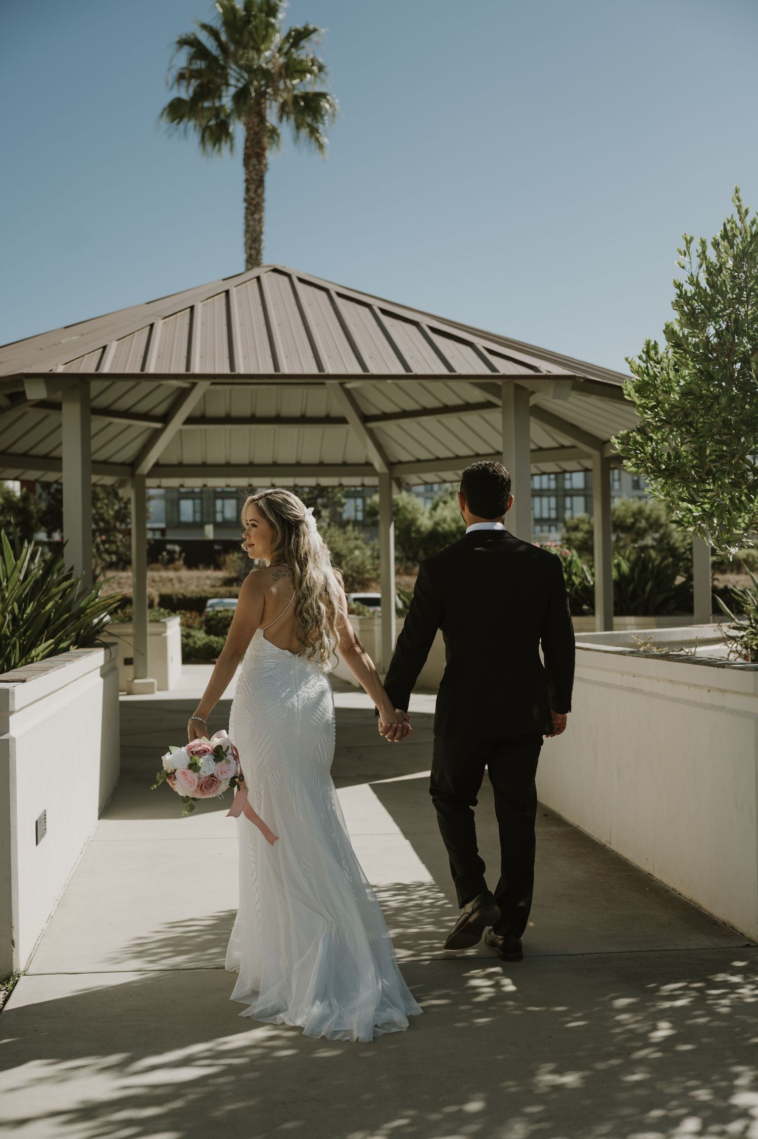 Newlyweds walk together under palm trees on a sunny outdoor pathway.