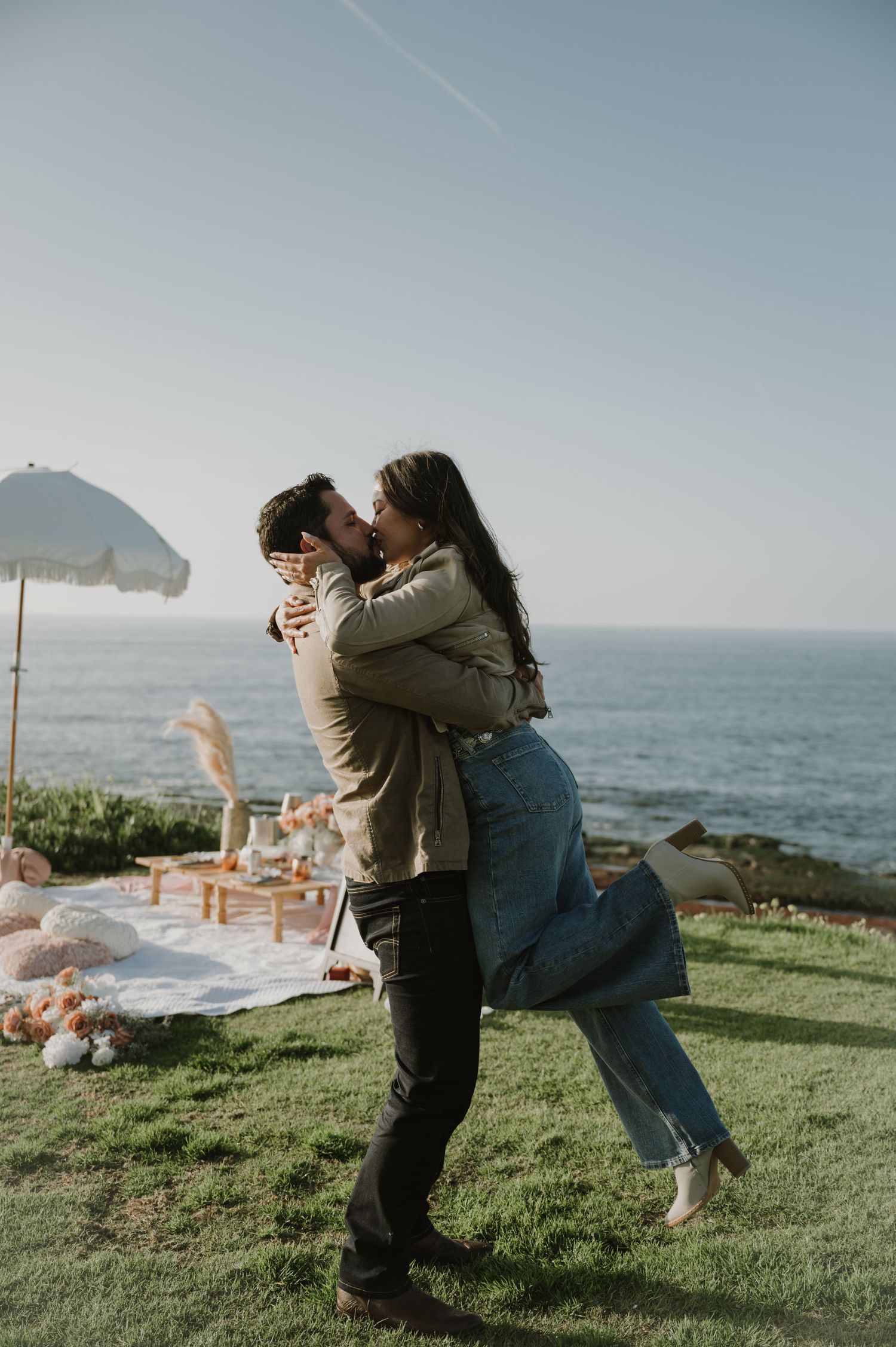 A couple celebrates their engagement on a scenic oceanside cliff with picnic setup.