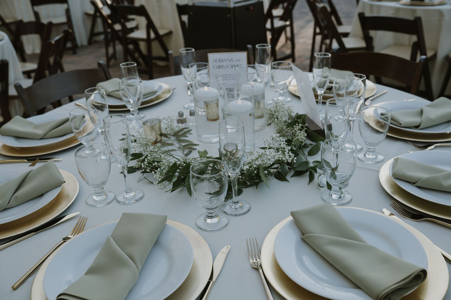 Elegant table settings with white linens, glass tableware, and sage green napkins at an outdoor wedding reception.