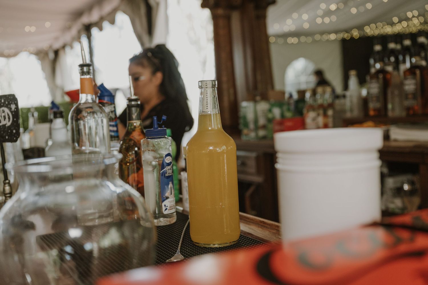 A bartender stands behind a rustic bar with bottles including an amber liquid-filled bottle at a outdoor event.