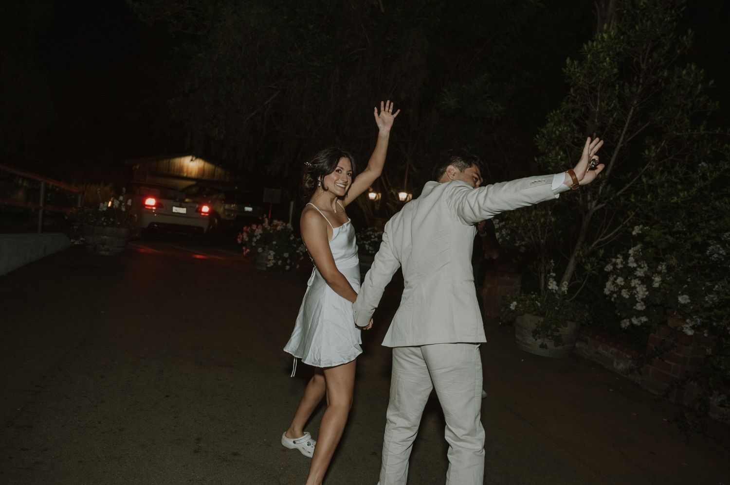 Bride and groom in white outfits dance joyfully together outdoors at night during wedding celebration.